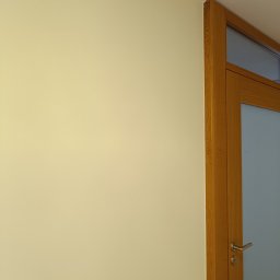 Matpol - Jasna, gładka ściana pomalowana na kremowo, przylegająca do drewnianej futryny drzwi z satynowym szkłem. Minimalistyczne wykończenie wnętrza.