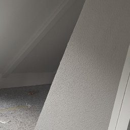 Matpol - Poddasze z oknem dachowym po remoncie: świeżo pomalowane ściany na biało z wyraźną fakturą, zabezpieczona podłoga, widoczne skosy i elementy konstrukcyjne.