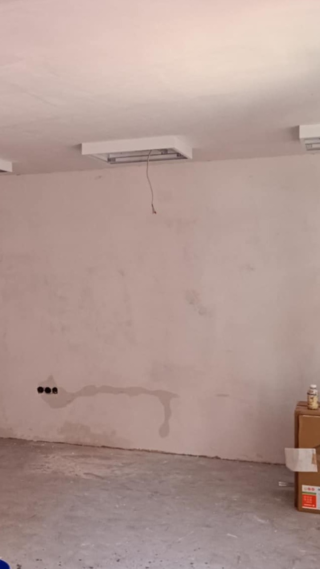 Ściana w trakcie szpachlowania z widocznymi śladami wilgoci i puszkami elektrycznymi, sufit z lampami. Podłoga betonowa. Widoczne kartony i butelka.