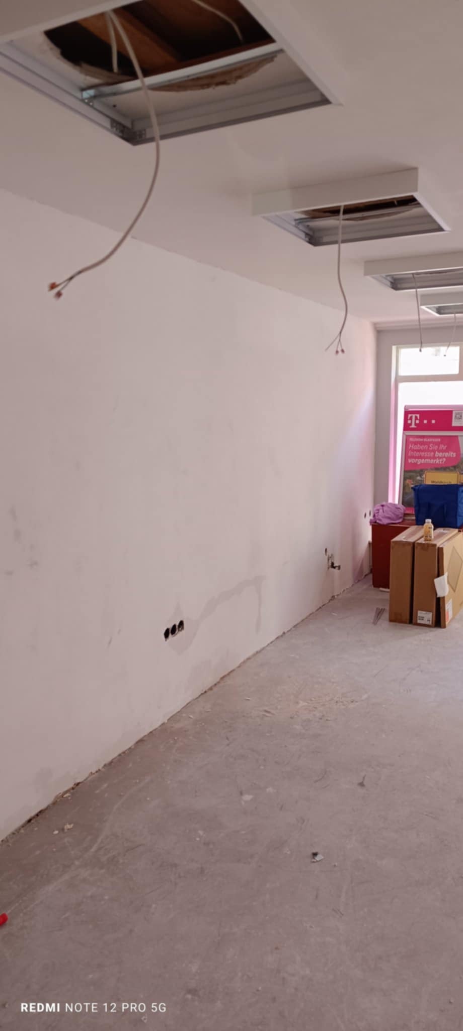 Biała ściana w trakcie szpachlowania z widocznymi przewodami, otwory w suficie pod instalację oświetleniową. Przy ścianie stoją kartony i inne przedmioty.