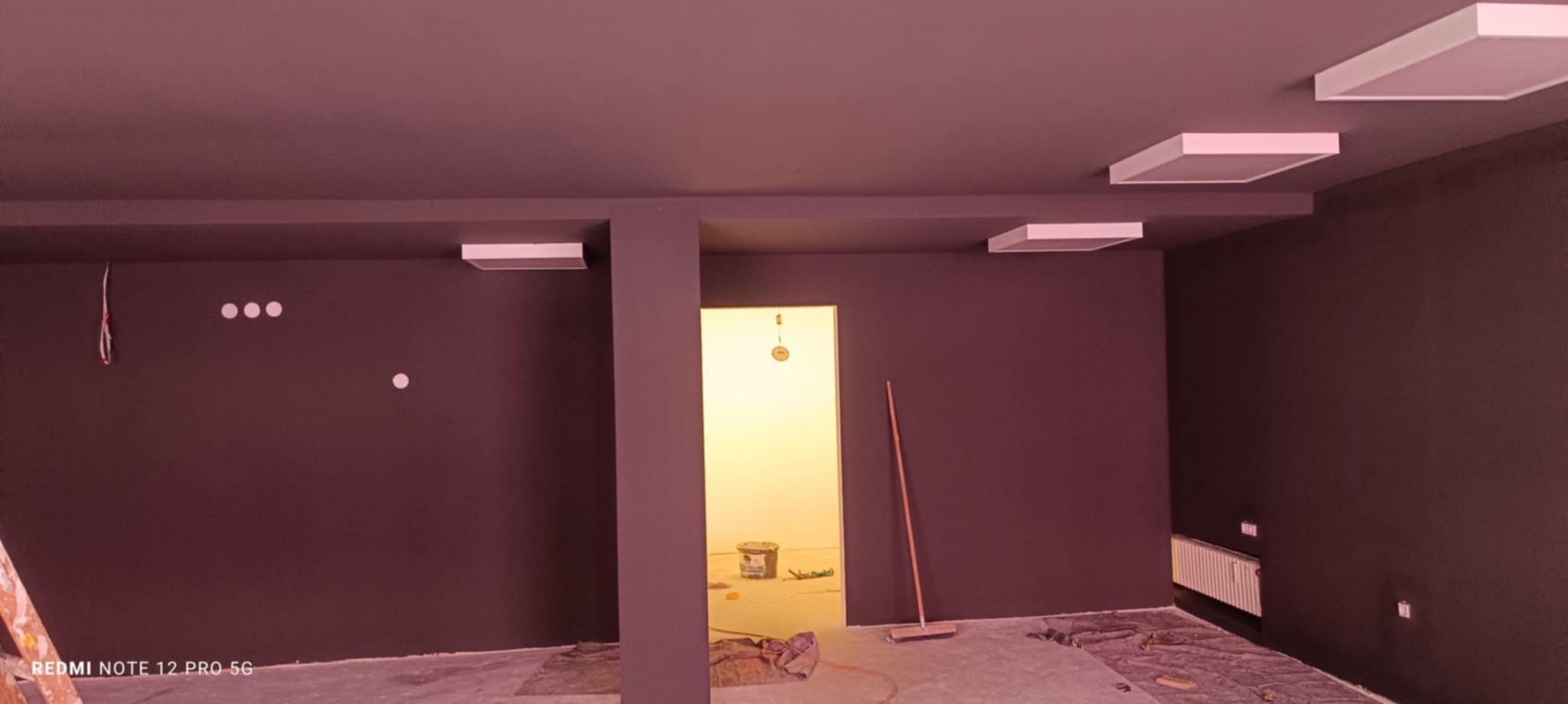 Pomieszczenie po malowaniu na ciemny kolor z białymi lampami sufitowymi. Widoczny sprzęt malarski i zakryta podłoga. W tle otwarte drzwi do innego pomieszczenia.