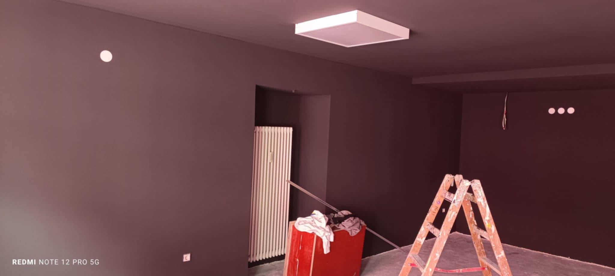 Pomieszczenie po malowaniu na ciemny kolor, widoczna drabina malarska, grzejnik i biała lampa sufitowa. Ściany jednolite, świeżo pomalowane.