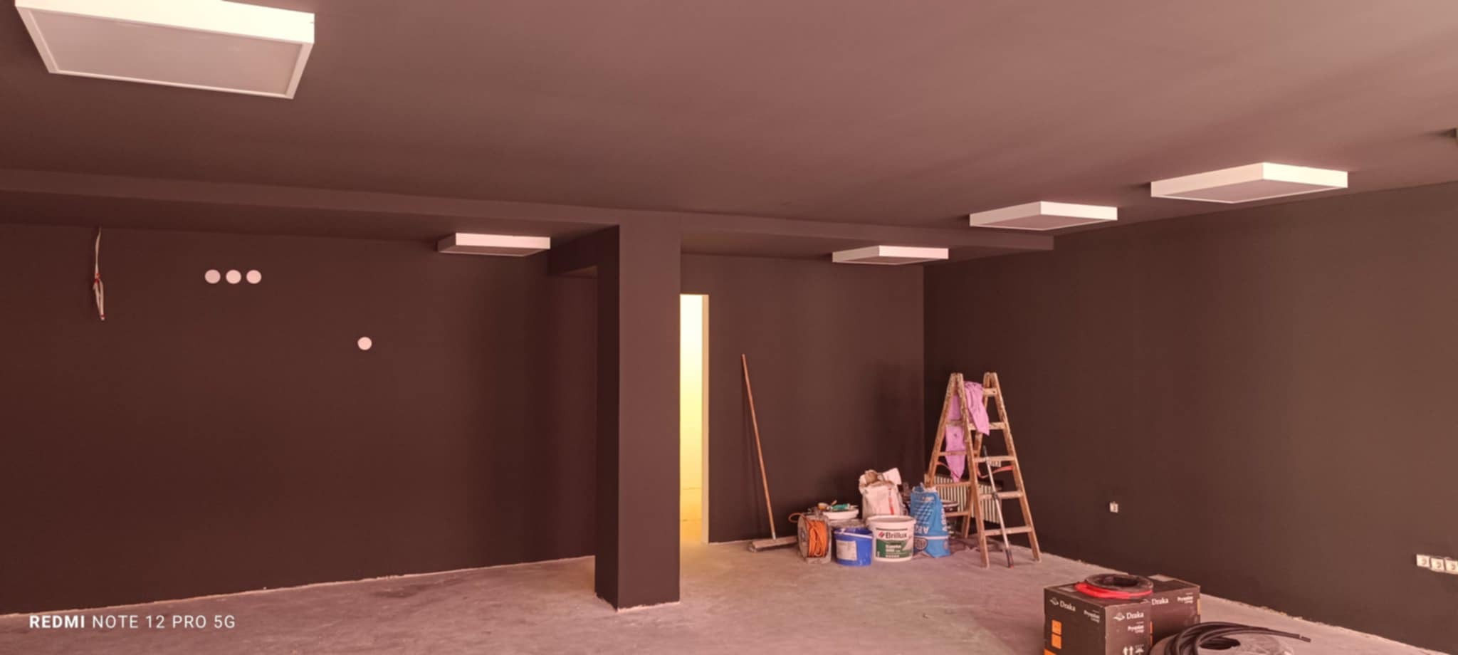 Pomieszczenie po malowaniu na brązowo z białymi lampami sufitowymi. Widoczne drabina, puszki z farbą i narzędzia malarskie. Świeżo pomalowane ściany i sufit.