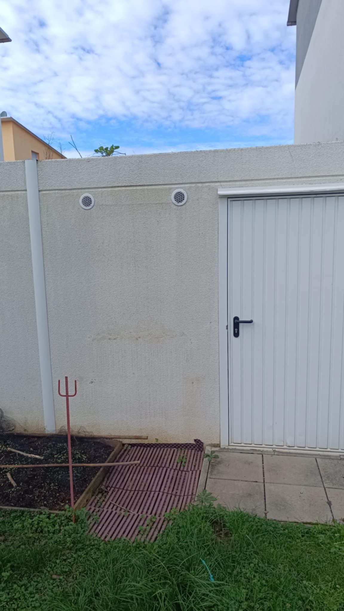 Szara, teksturowana ściana zewnętrzna z dwoma okrągłymi otworami wentylacyjnymi i białymi drzwiami garażowymi, widoczne narzędzie ogrodnicze i fragment grządki.