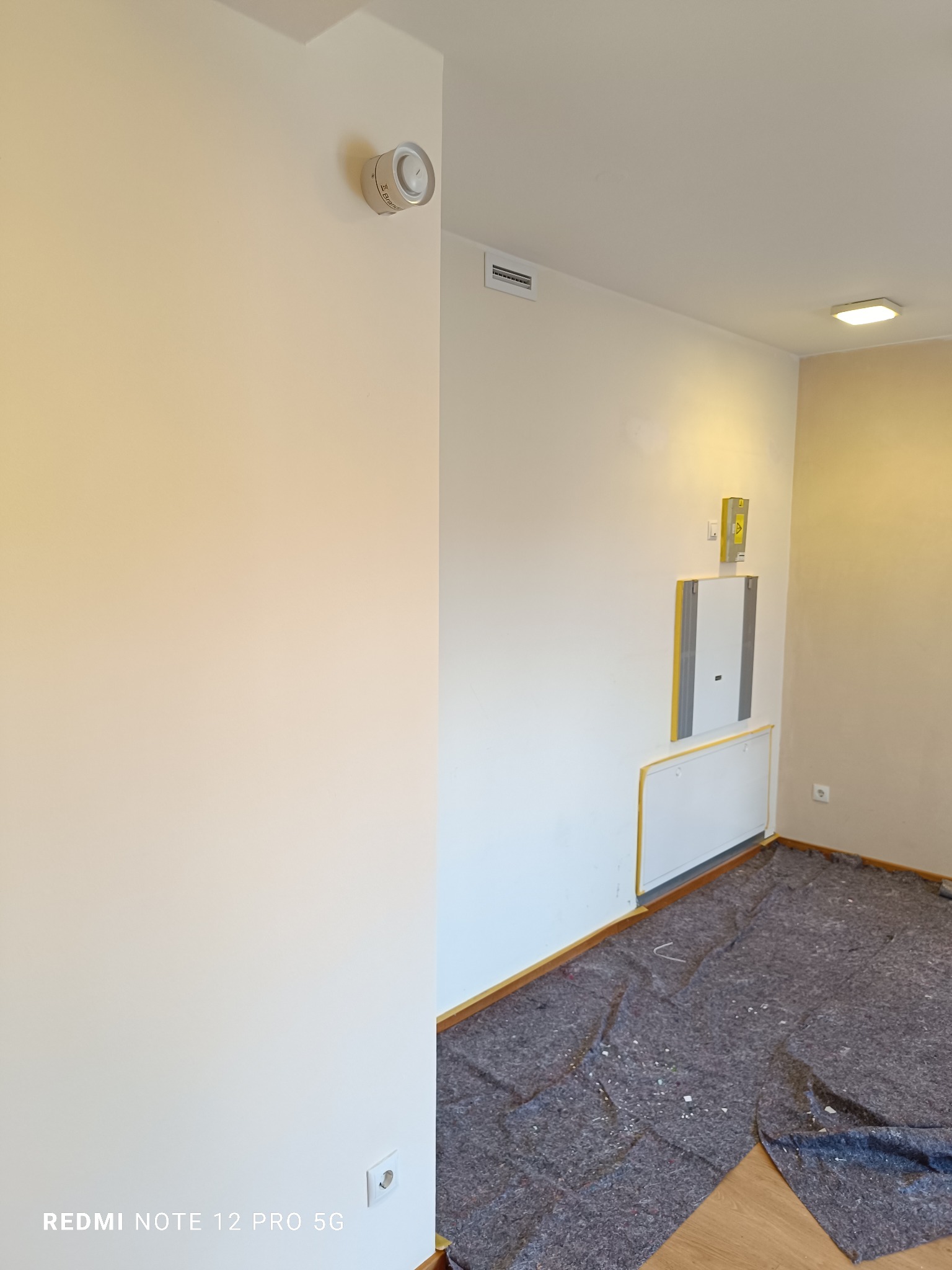 Jasny pokój w trakcie remontu: świeżo pomalowane ściany w odcieniach beżu, zabezpieczona podłoga, widoczny czujnik dymu i skrzynka elektryczna.