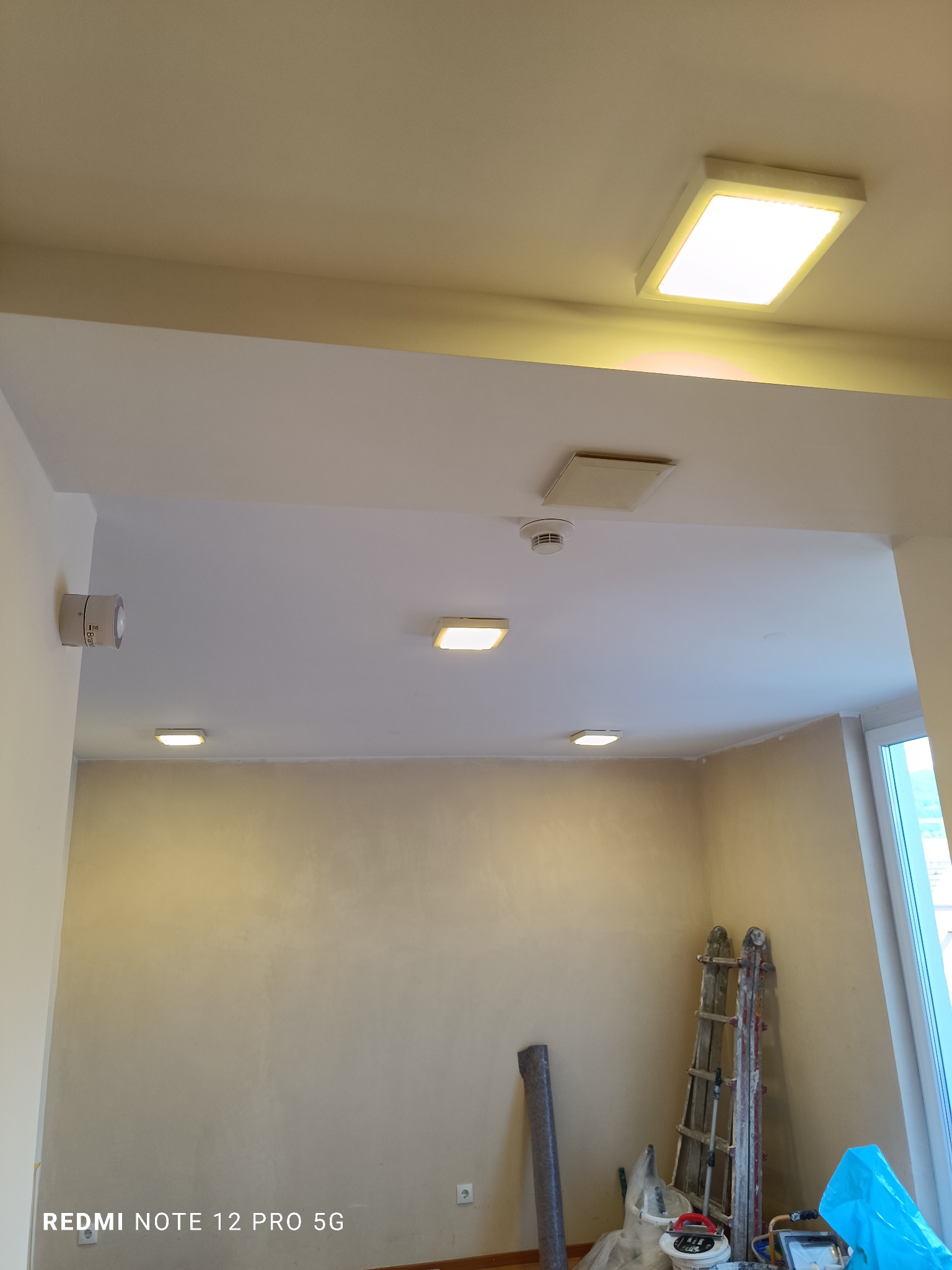 Wnętrze pokoju po szpachlowaniu, widoczne narzędzia malarskie, drabina, sufit z oświetleniem LED. Ściany przygotowane do malowania, neutralny odcień, okno w tle.