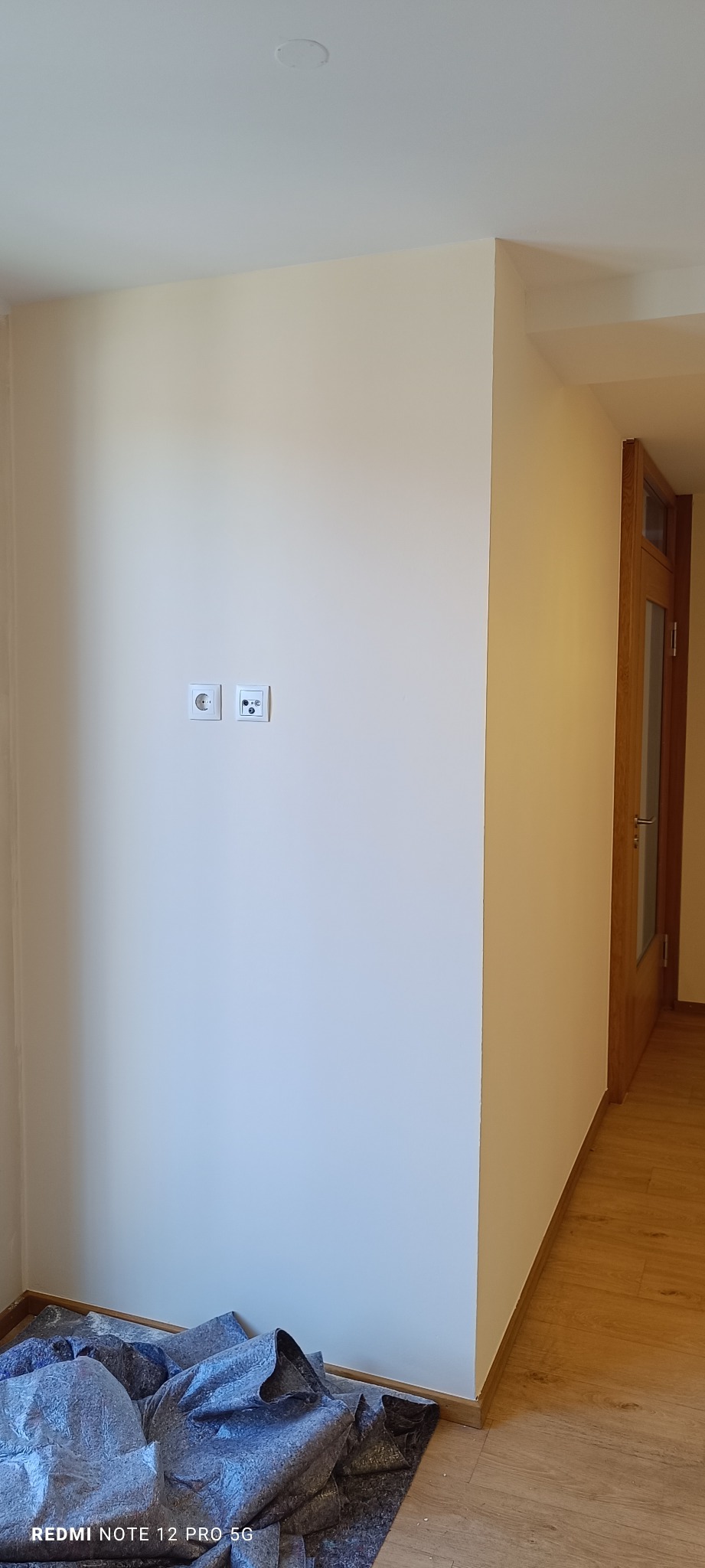 Jasna ściana po świeżym malowaniu, z widocznymi gniazdkami elektrycznymi i zabezpieczeniem podłogi. Fragment drewnianej podłogi i drzwi w tle. Neutralne oświetlenie.