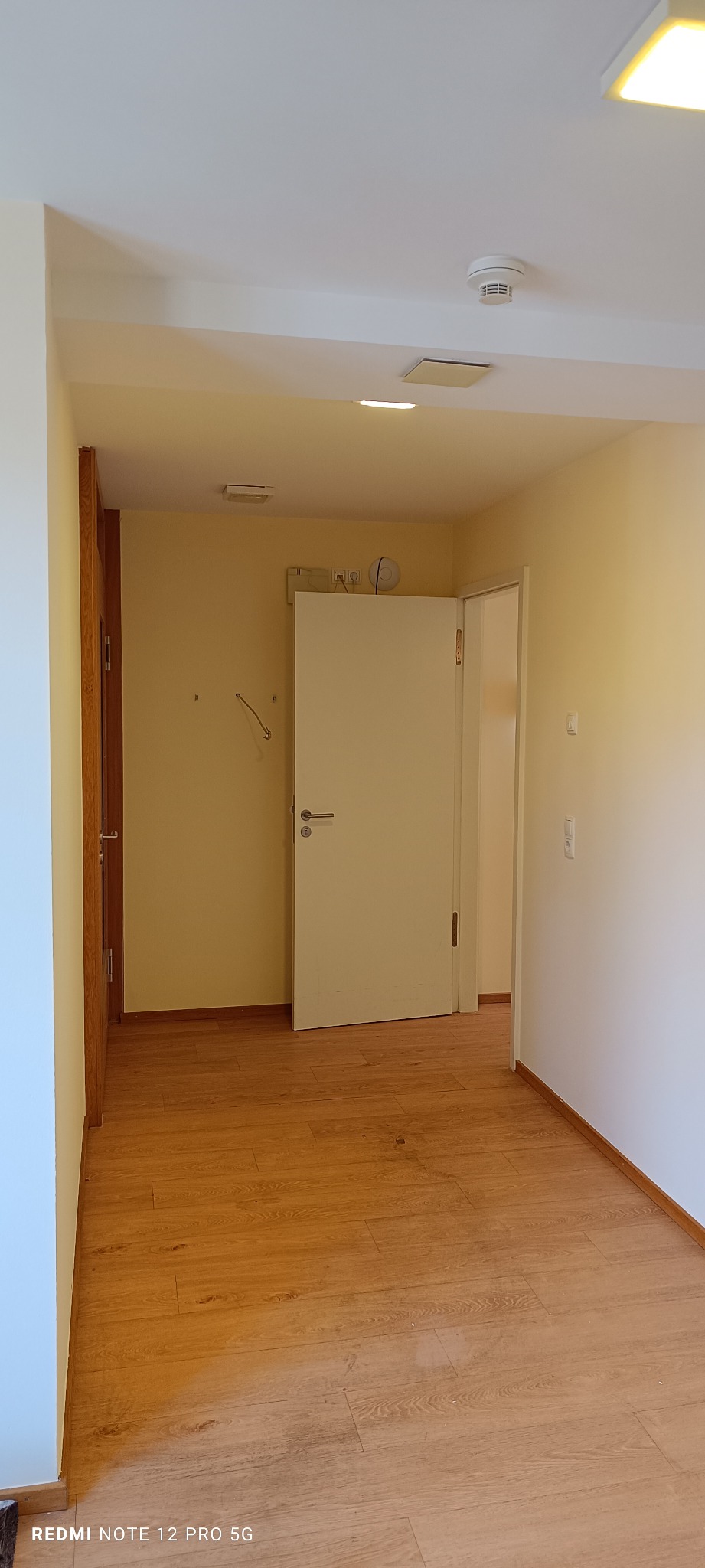 Jasne wnętrze z drewnianą podłogą i białą ścianą. Widoczne drzwi, włączniki i gniazdka elektryczne. Minimalistyczny design, ciepłe oświetlenie sufitowe.