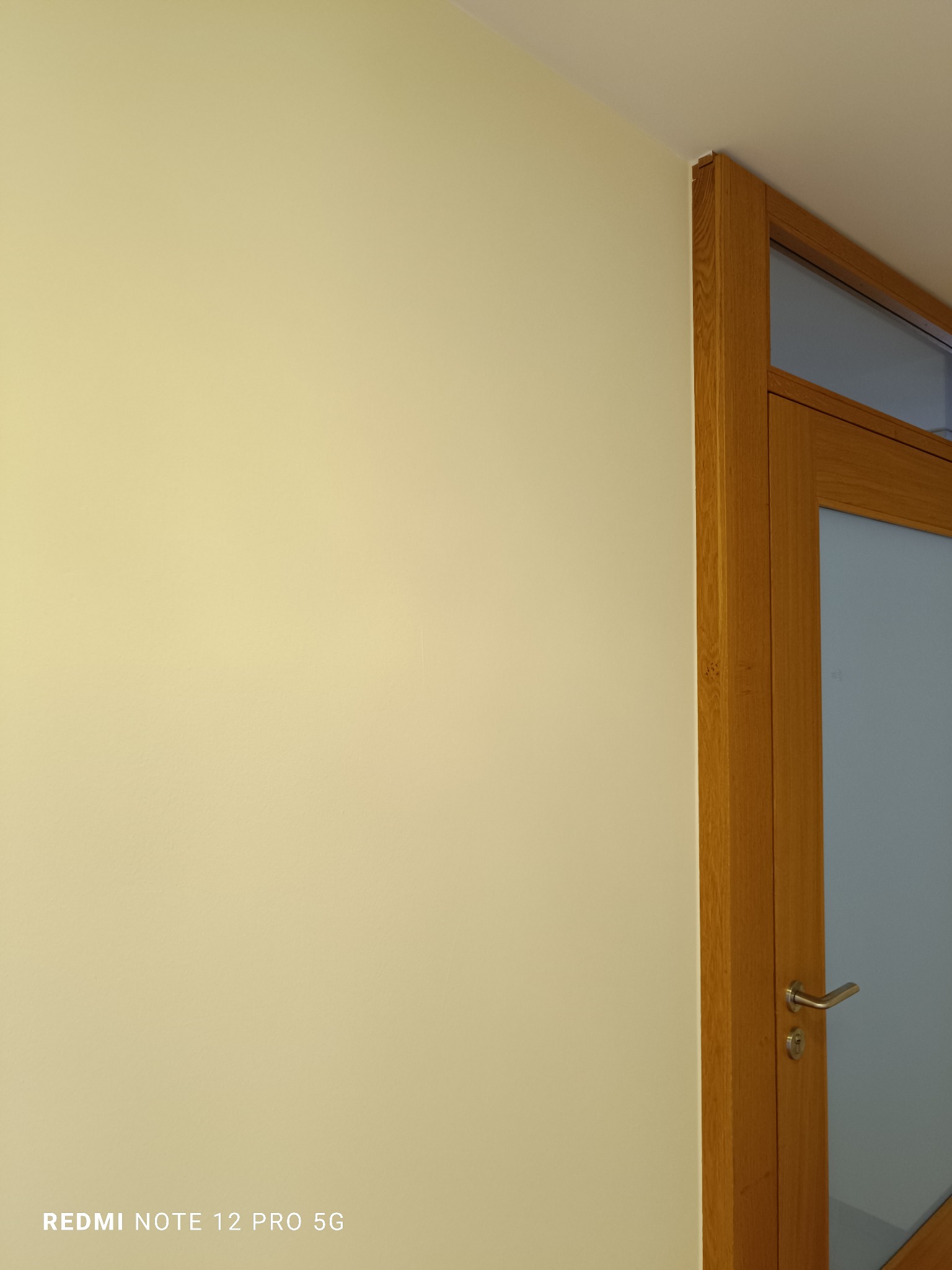 Jasna, gładka ściana pomalowana na kremowo, przylegająca do drewnianej futryny drzwi z satynowym szkłem. Minimalistyczne wykończenie wnętrza.