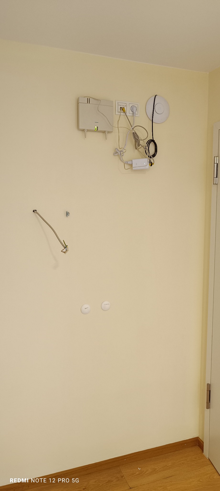 Beżowa ściana z wystającymi kablami, gniazdkami i routerem. Widoczne dwa zaślepione otwory w ścianie. Podłoga drewniana.