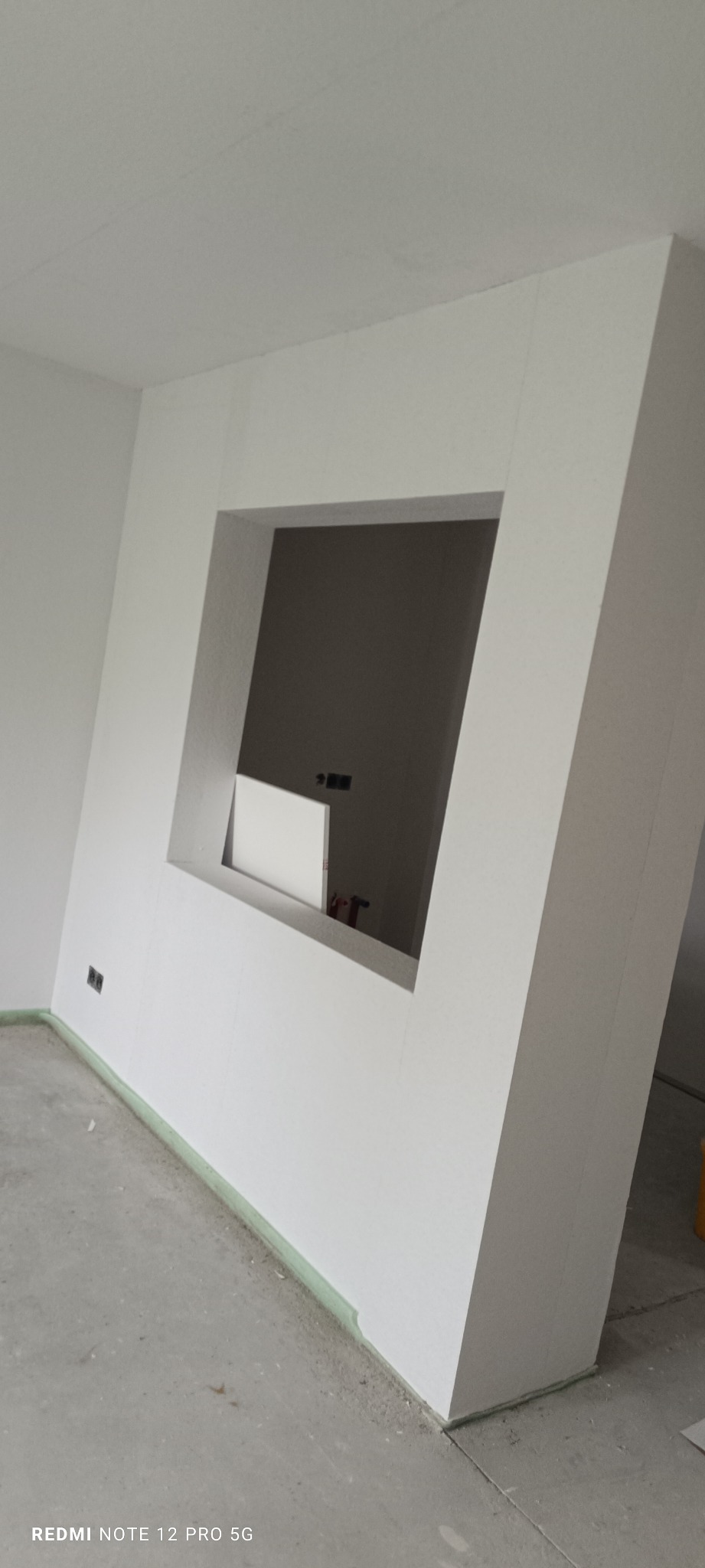 Biała ściana z asymetrycznym otworem w nowym mieszkaniu, widoczne gniazdka elektryczne i zabezpieczenie podłogi. Ściany przygotowane do malowania.