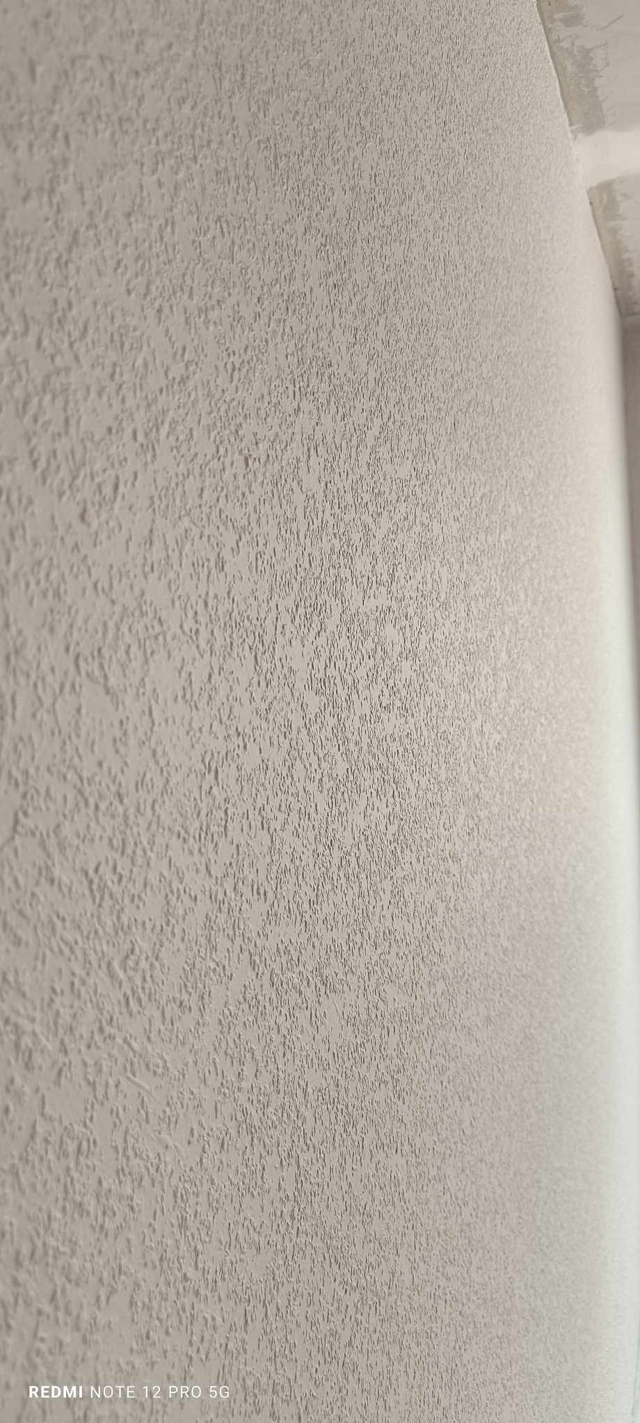 Ściana pokryta jasną, strukturalną gładzią o wyraźnej fakturze, widoczna pod kątem. Wykończenie ściany w odcieniach beżu i szarości, z delikatnym cieniowaniem.