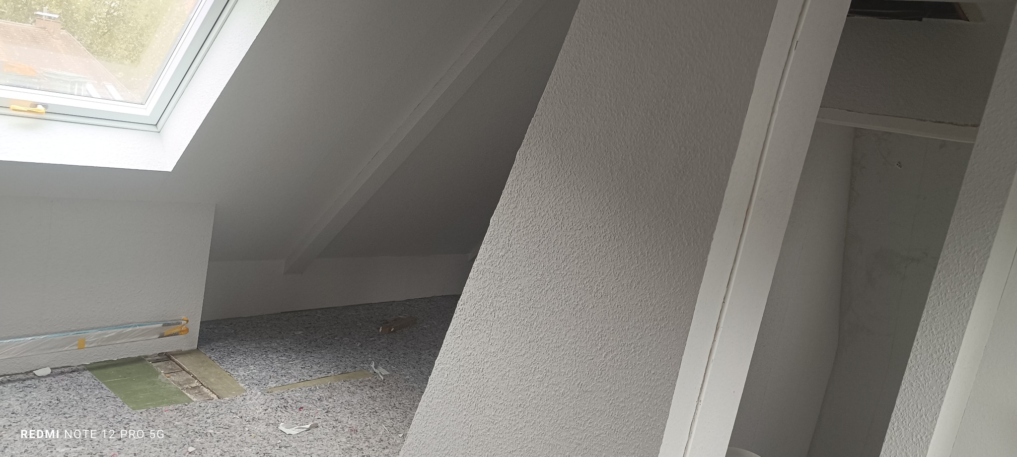 Poddasze z oknem dachowym po remoncie: świeżo pomalowane ściany na biało z wyraźną fakturą, zabezpieczona podłoga, widoczne skosy i elementy konstrukcyjne.