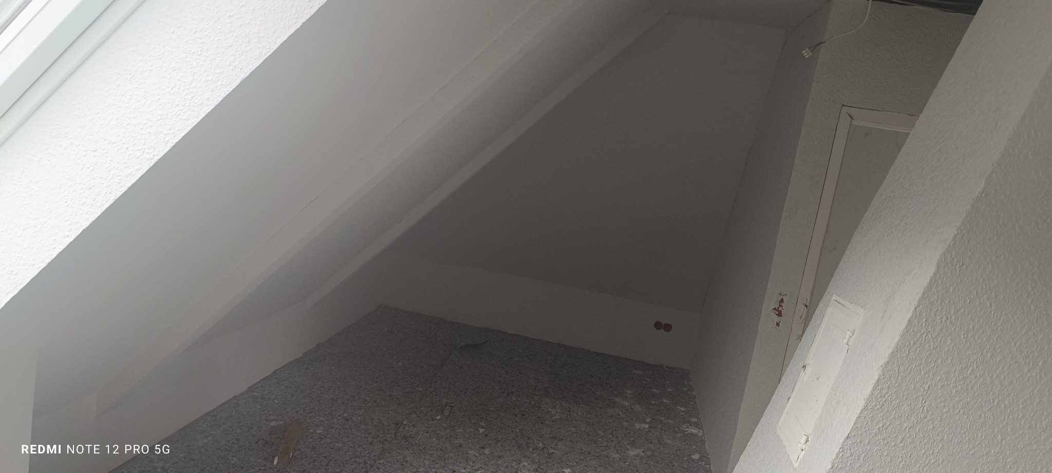 Poddasze po remoncie. Ściany i skosy pomalowane na biało, widoczne gniazdka elektryczne. Na podłodze resztki po remoncie. Ujęcie z dołu.