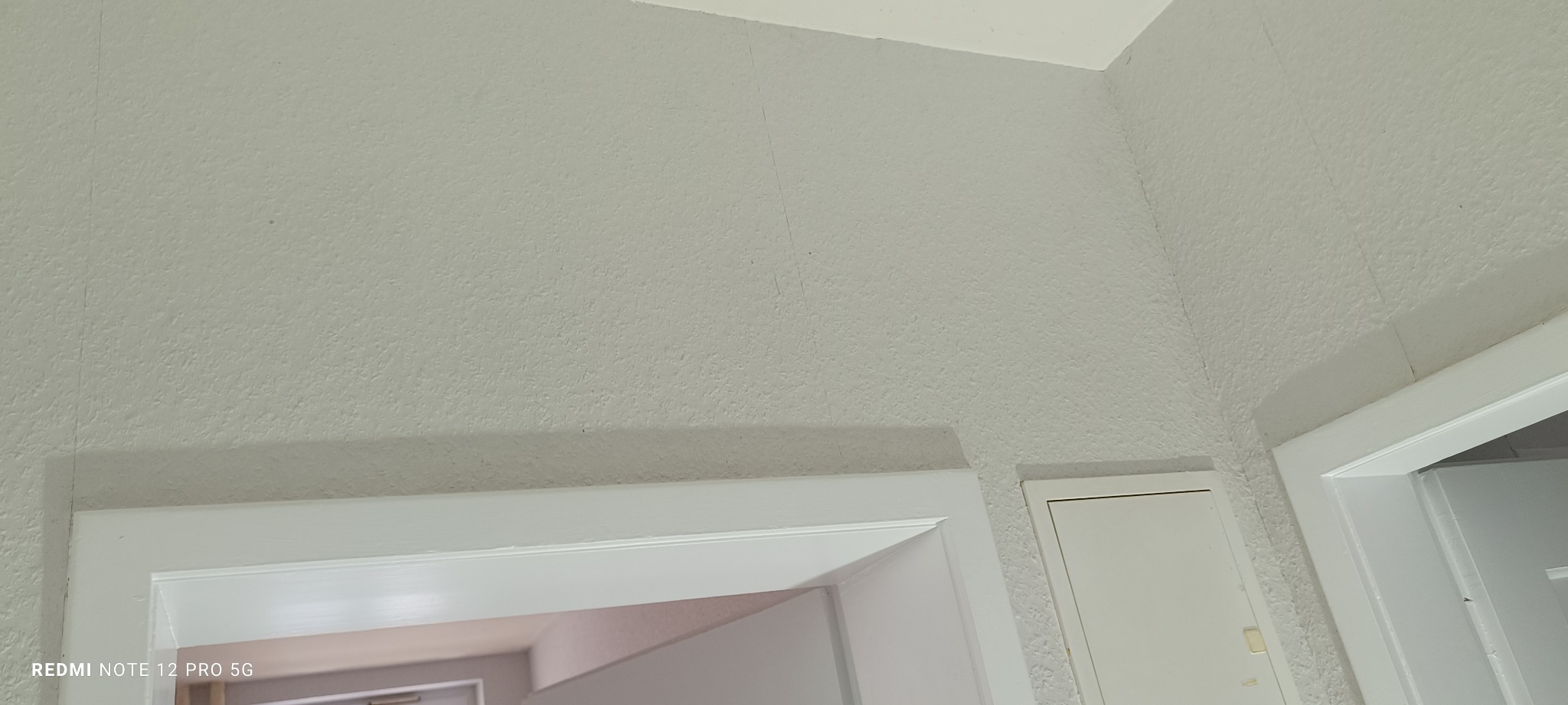 Ściana z fakturą pomalowana na biało, widoczny fragment ościeżnicy drzwi. Delikatne światło podkreśla strukturę. Widoczne minimalne niedoskonałości.