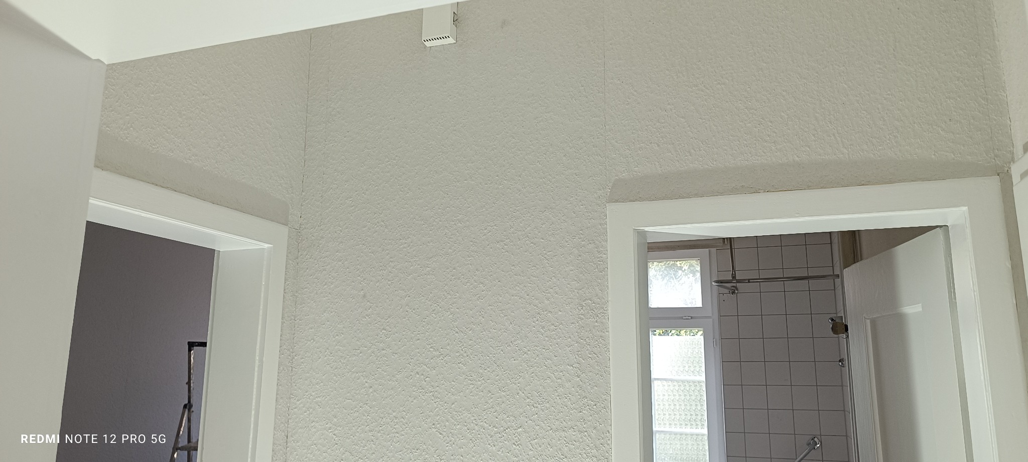 Biała ściana z fakturą, widoczne dwa białe futryny drzwi, w tle drabina malarska i łazienka z białymi płytkami. Świeżo pomalowane ściany z widoczną strukturą.