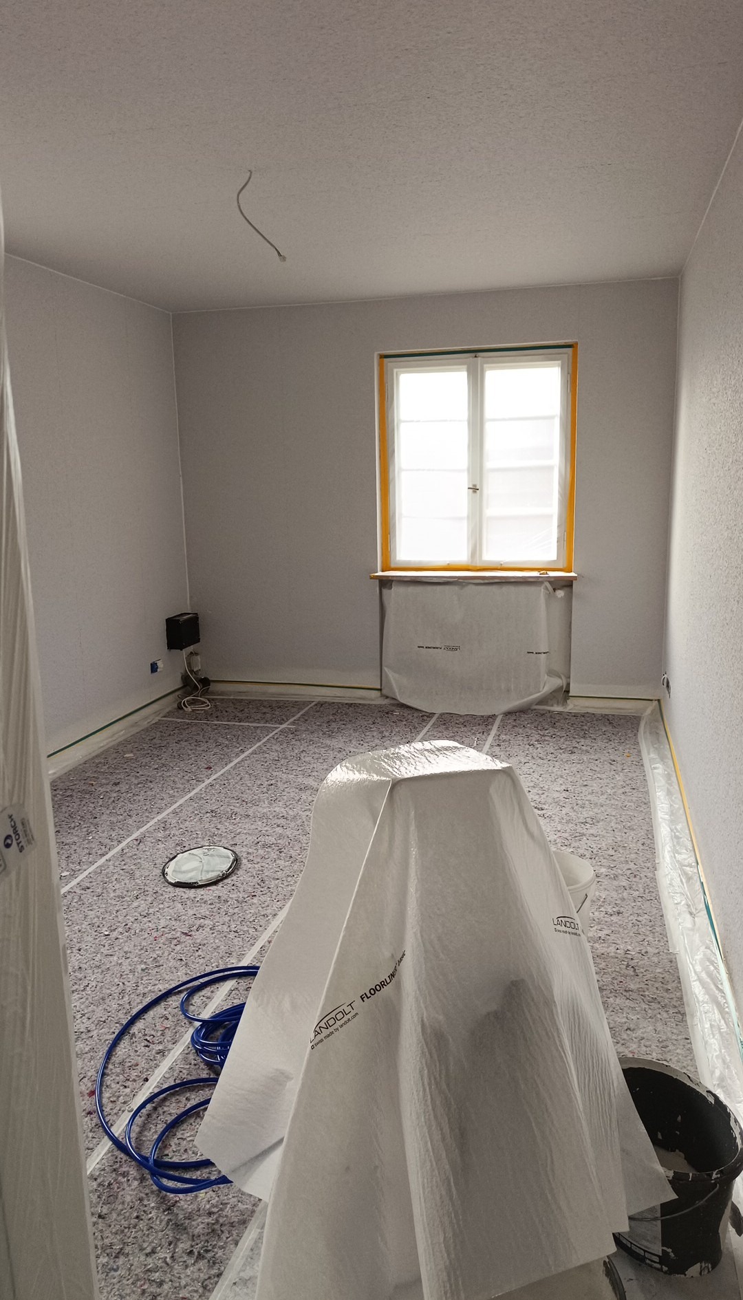 Pokój w trakcie przygotowań do prac wykończeniowych. Zabezpieczone podłogi i okna, widoczne narzędzia malarskie.