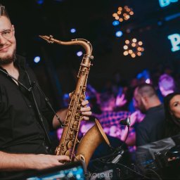 ART Jonasz Styszyński - Saksofonista w okularach, grający na saksofonie w klubie. W tle publiczność i neonowy napis 'POINT CLUB'.