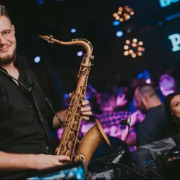 Saksofonista w okularach, grający na saksofonie w klubie. W tle publiczność i neonowy napis 'POINT CLUB'.