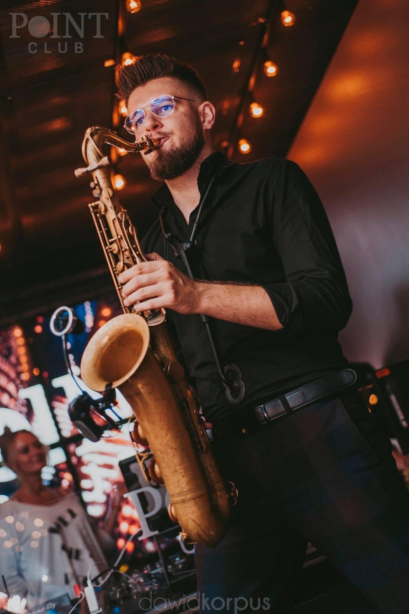 Saksofonista z brodą i okularami, w czarnej koszuli, grający na saksofonie na tle klubu z dekoracyjnymi światłami, częściowo widoczna publiczność i DJ.