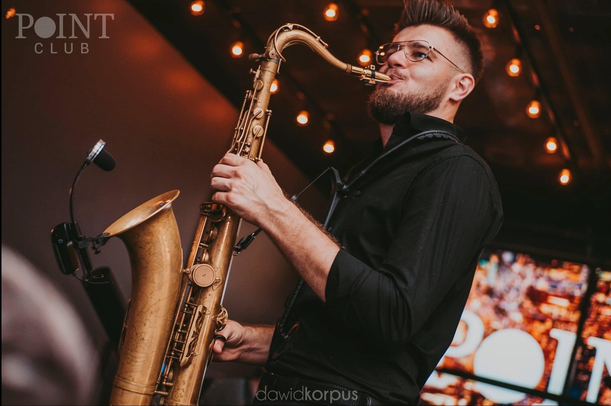 Saksofonista w czarnej koszuli gra na saksofonie w klubie z oświetleniem, widoczny mikrofon i logo klubu w tle, artysta z brodą i okularami.