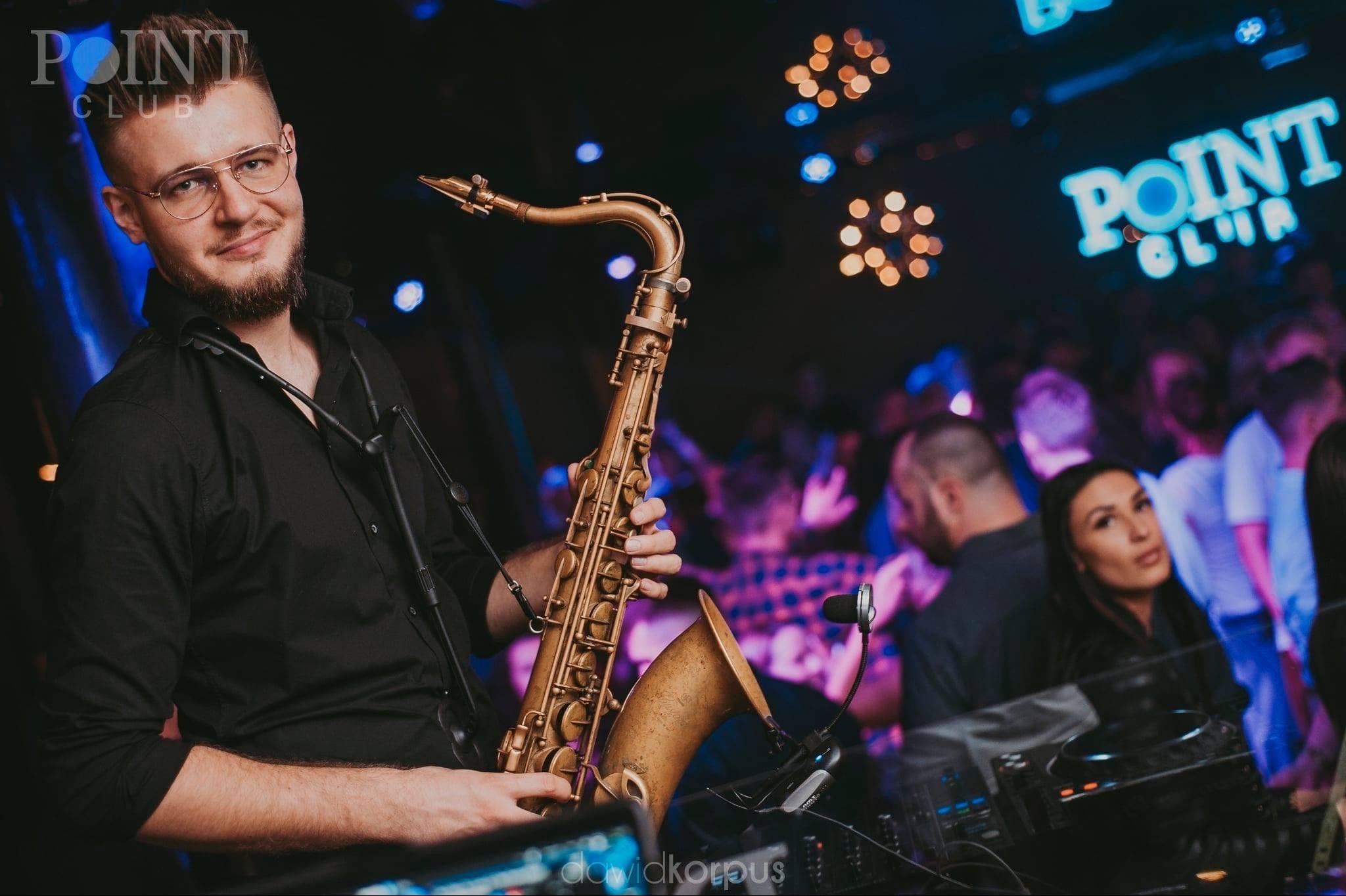 Saksofonista w okularach, grający na saksofonie w klubie. W tle publiczność i neonowy napis 'POINT CLUB'.