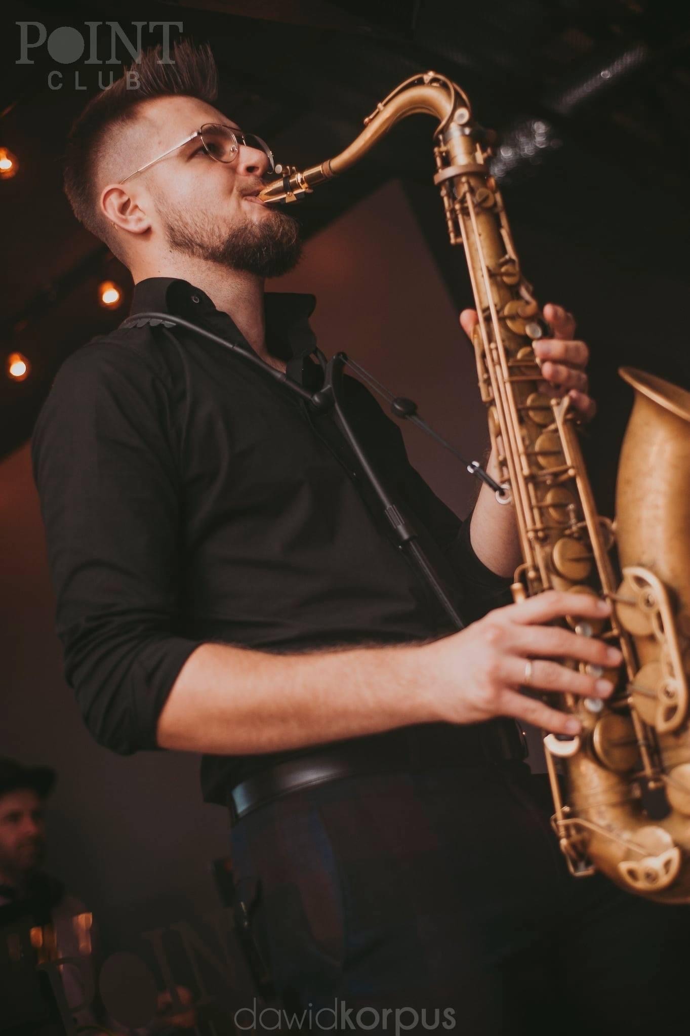 Saksofonista w czarnej koszuli gra w Obornikach. Artysta z brodą i okularach, w tle światła i perkusja. Klimatyczne ujęcie muzyka podczas występu na żywo.