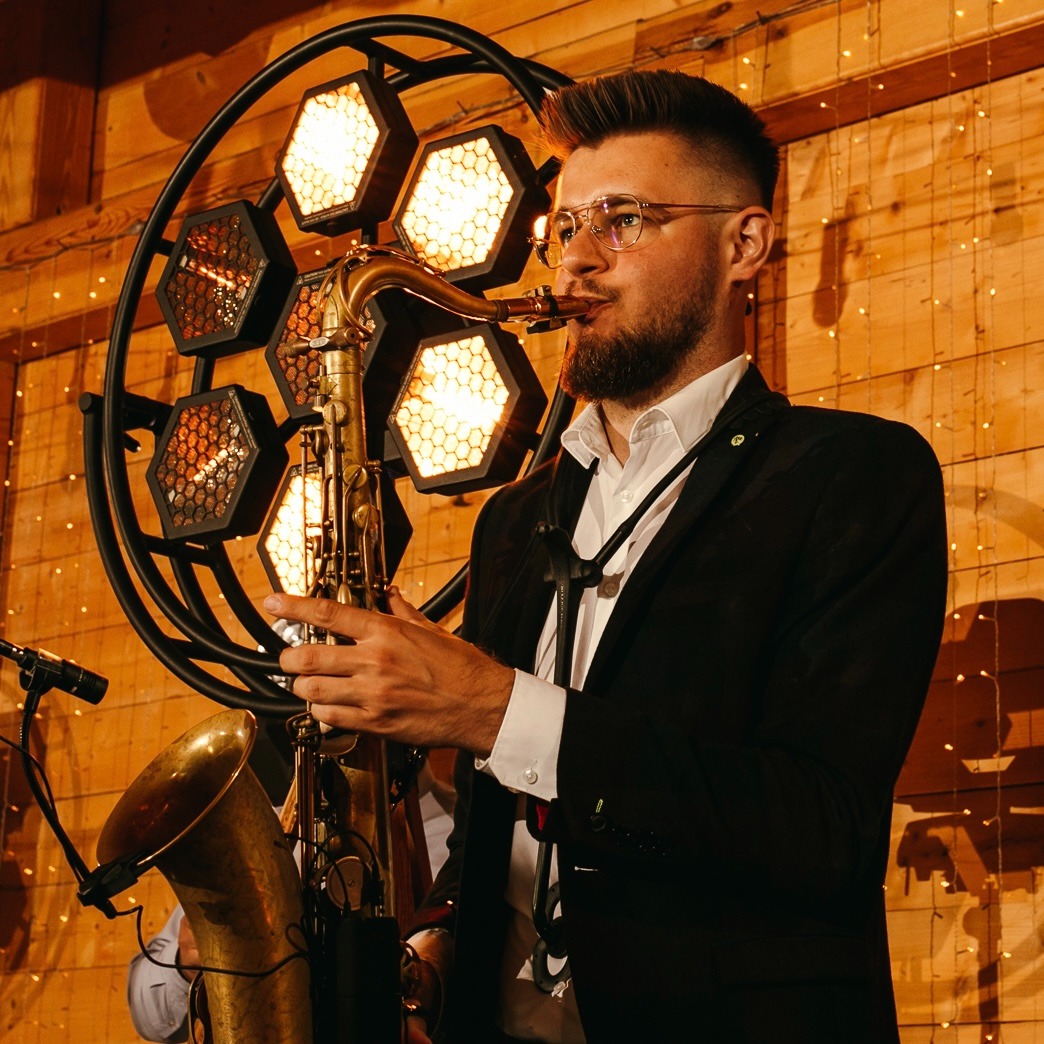 Saksofonista w czarnym garniturze gra na saksofonie na tle drewnianej ściany z dekoracyjnym oświetleniem. Widoczne detale instrumentu i profesjonalny wygląd muzyka.