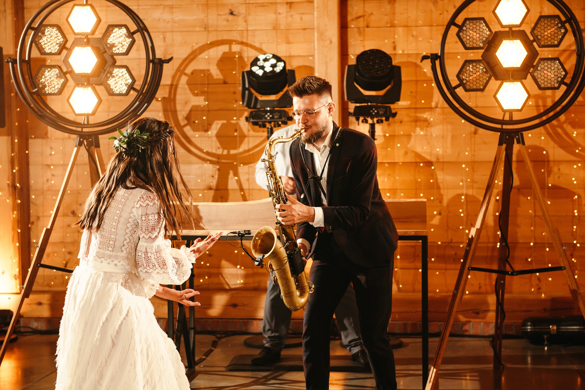 Saksofonista w bordowym garniturze gra na saksofonie podczas wesela, panna młoda tańczy w białej sukni, drewniane tło z dekoracyjnymi światłami.
