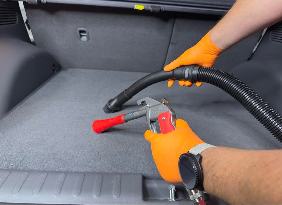 Czyszczenie tapicerki bagażnika samochodowego za pomocą odkurzacza i specjalistycznego pistoletu z czerwoną końcówką, wykonywane przez osobę w pomarańczowych rękawicach.