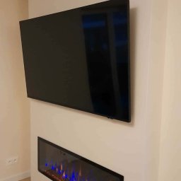 Milimetr Kwadrat - Remonty i wykończenia Piotr Stróżewski - Nowoczesny salon z zamontowanym telewizorem i podłużnym kominkiem elektrycznym z niebiesko-pomarańczowymi płomieniami, w minimalistycznym wnętrzu.