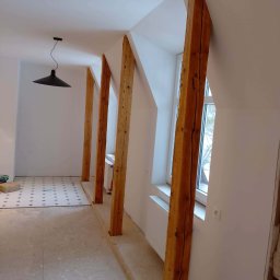 Milimetr Kwadrat - Remonty i wykończenia Piotr Stróżewski - Wnętrze poddasza w trakcie remontu: drewniane belki konstrukcyjne, białe ściany, okna i fragment podłogi z wzorem. Widoczny etap prac wykończeniowych.