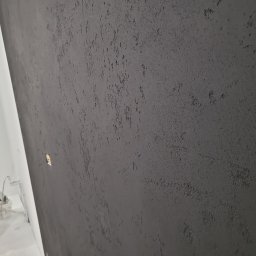 Milimetr Kwadrat - Remonty i wykończenia Piotr Stróżewski - Szara ściana z dekoracyjną strukturą, widoczna pod kątem. W tle biała ściana i sprzęt malarski. Efektowna faktura na ścianie.