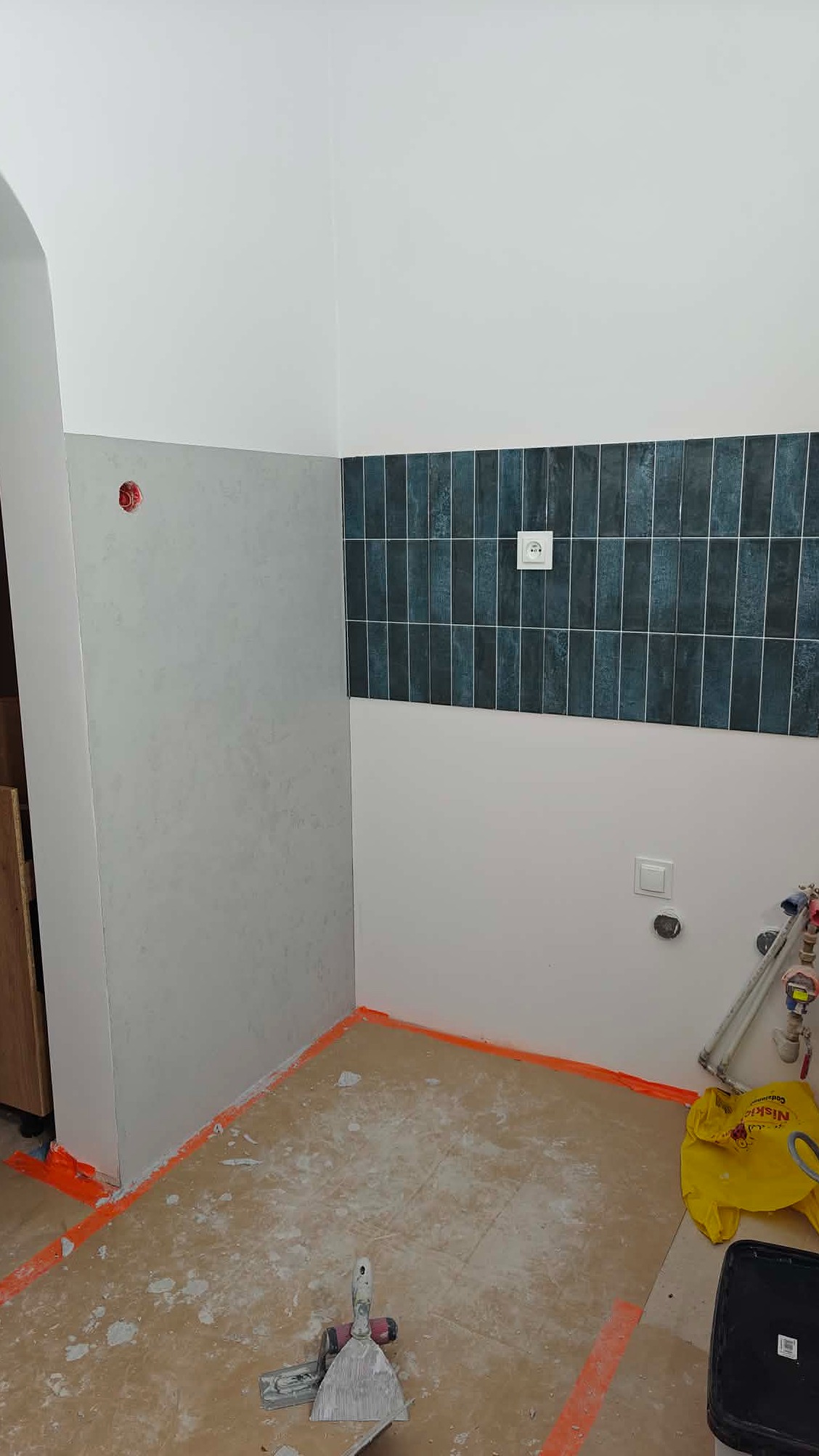 Fragment wnętrza w trakcie prac wykończeniowych: położone płytki na ścianie, przygotowane ściany do malowania, zabezpieczona podłoga, narzędzia pracy.