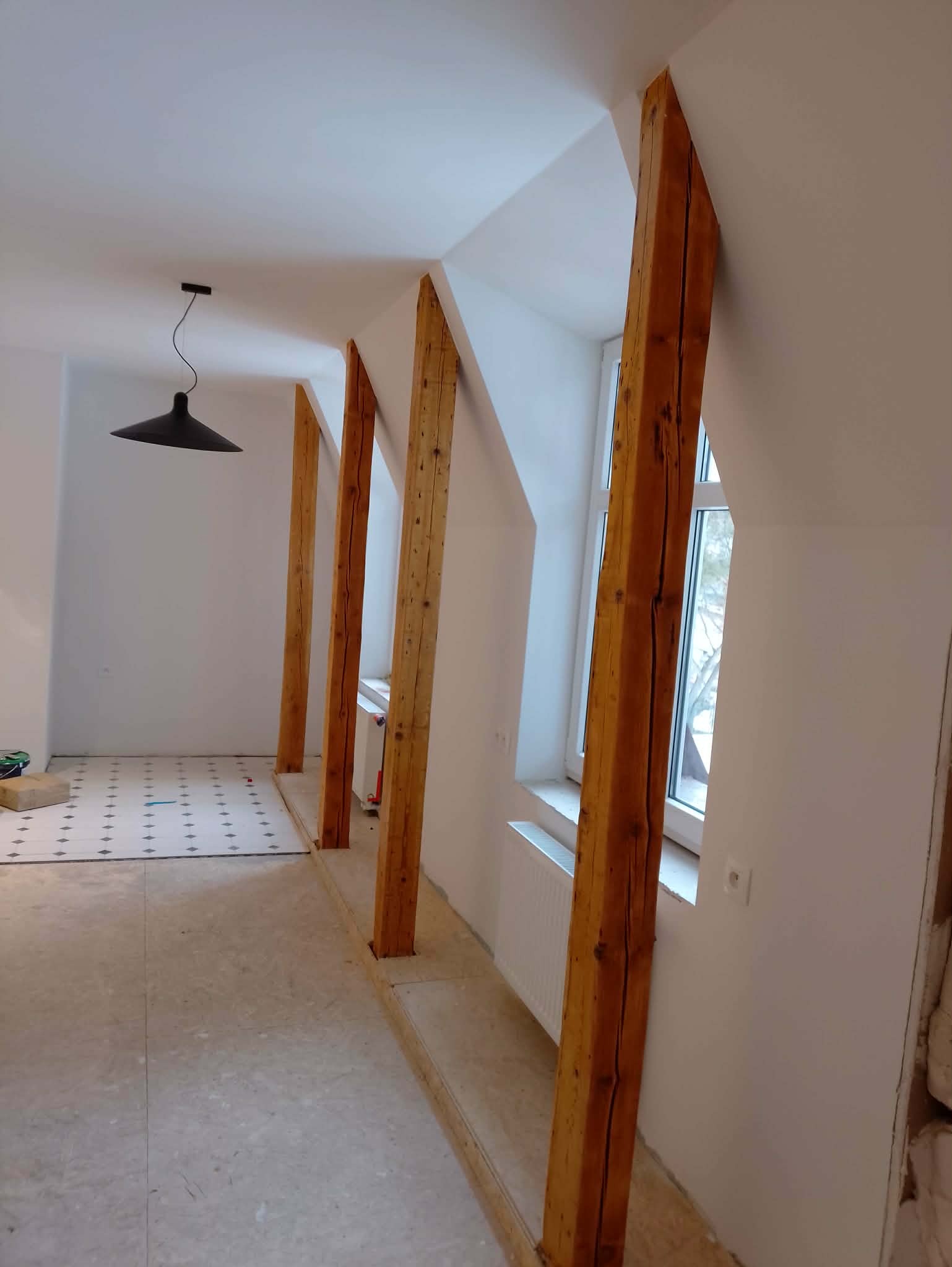 Wnętrze poddasza w trakcie remontu: drewniane belki konstrukcyjne, białe ściany, okna i fragment podłogi z wzorem. Widoczny etap prac wykończeniowych.