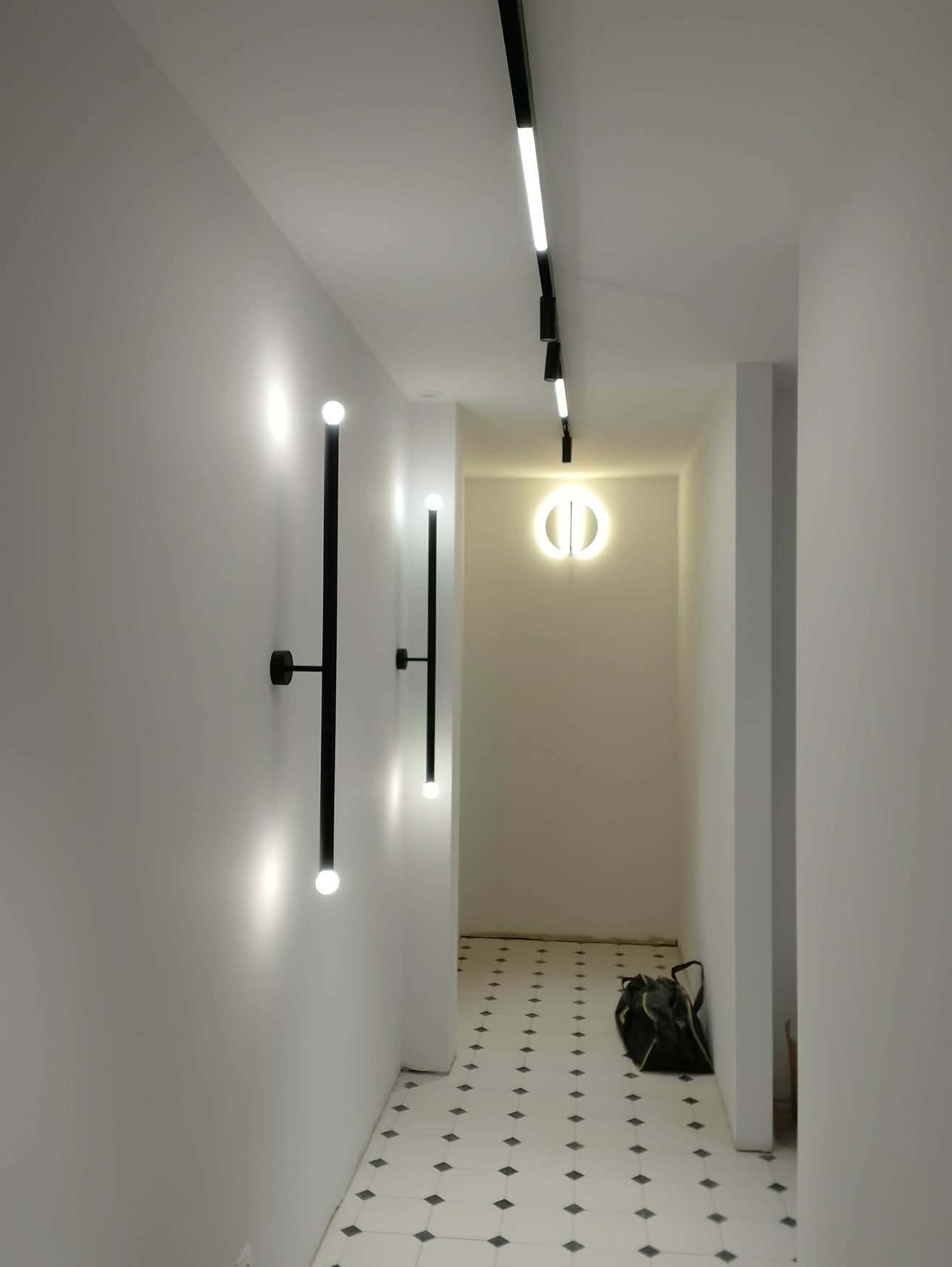 Nowoczesny korytarz z oświetleniem LED: liniowe na suficie, pionowe kinkiety na ścianach i okrągła lampa na końcu. Białe ściany i podłoga w czarno-białą szachownicę.