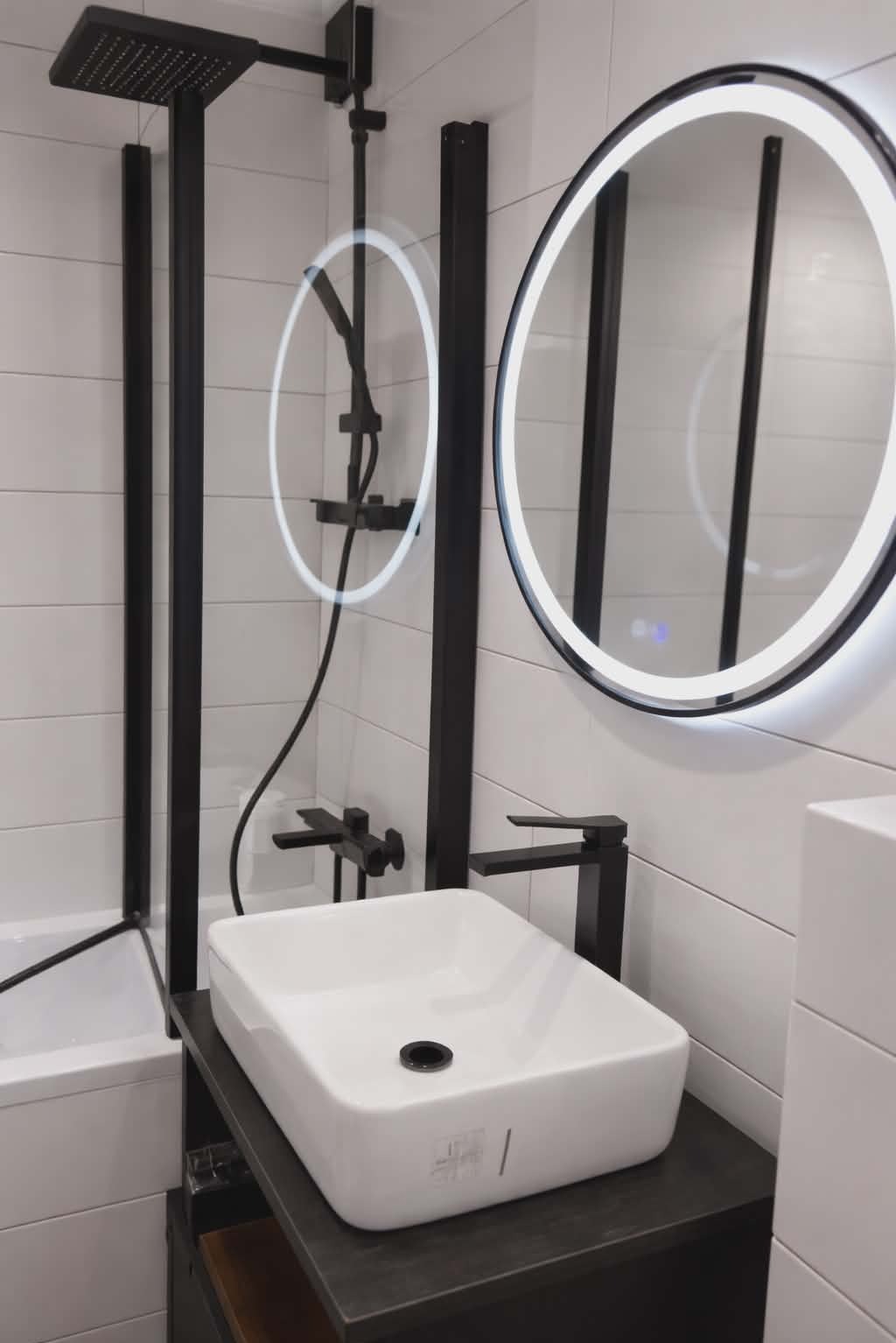 Nowoczesna łazienka z białą umywalką nablatową, czarną armaturą i okrągłym lustrem z podświetleniem LED. Minimalistyczny design z płytkami imitującymi drewno.