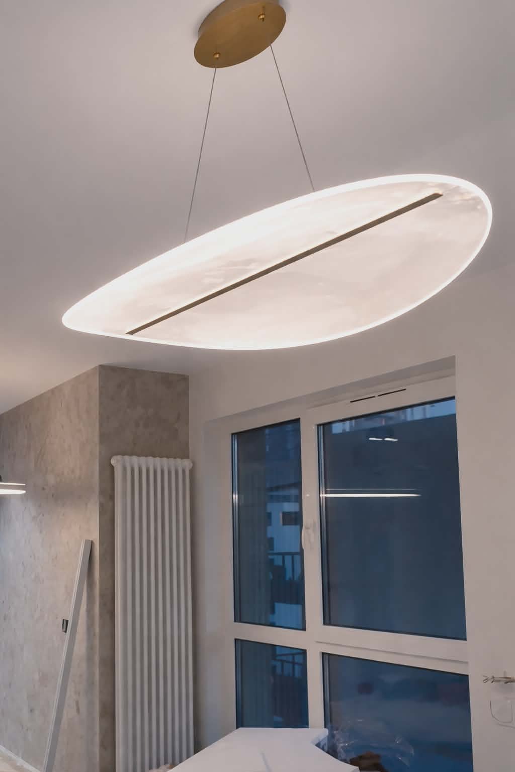 Nowoczesna lampa sufitowa w kształcie elipsy z czarnym akcentem, zawieszona w jasnym wnętrzu z oknem i grzejnikiem. Minimalistyczny design oświetlenia.