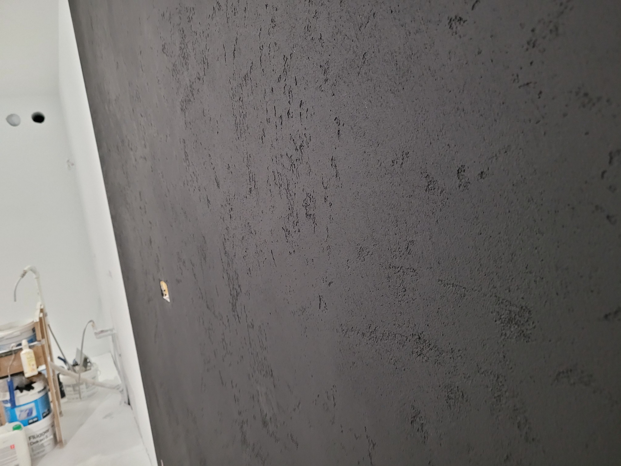 Szara ściana z dekoracyjną strukturą, widoczna pod kątem. W tle biała ściana i sprzęt malarski. Efektowna faktura na ścianie.