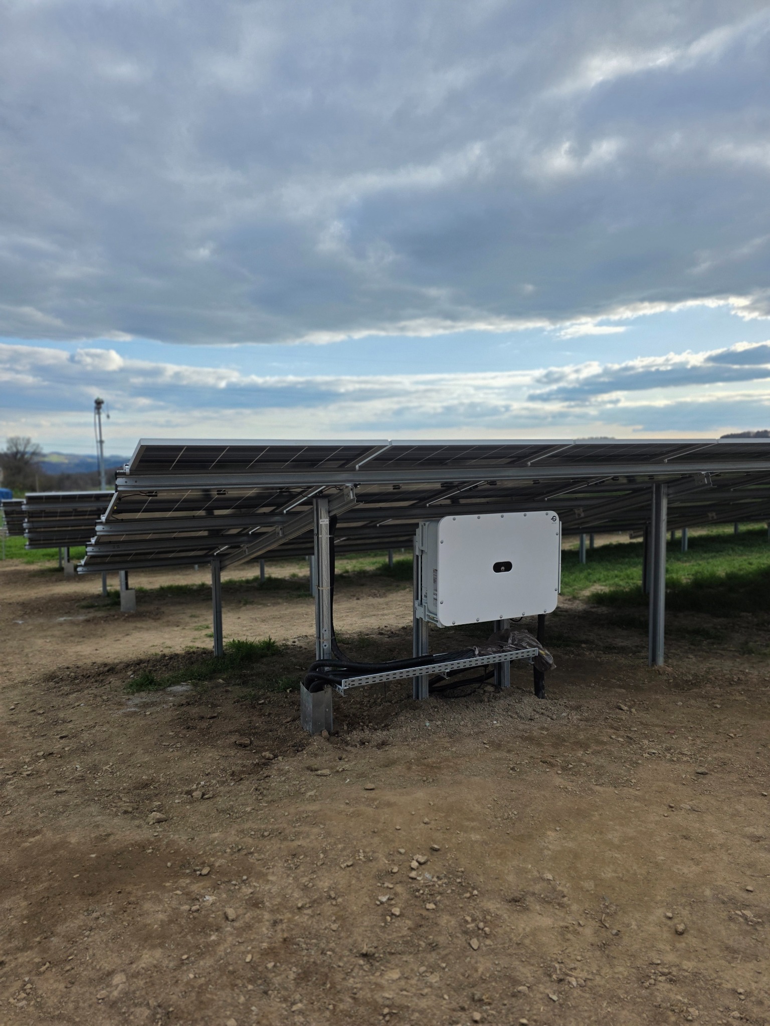 Instalacja paneli słonecznych na metalowej konstrukcji z białym inwerterem na tle pochmurnego nieba i zielonego krajobrazu. Widok z dołu, podkreślający solidność montażu.