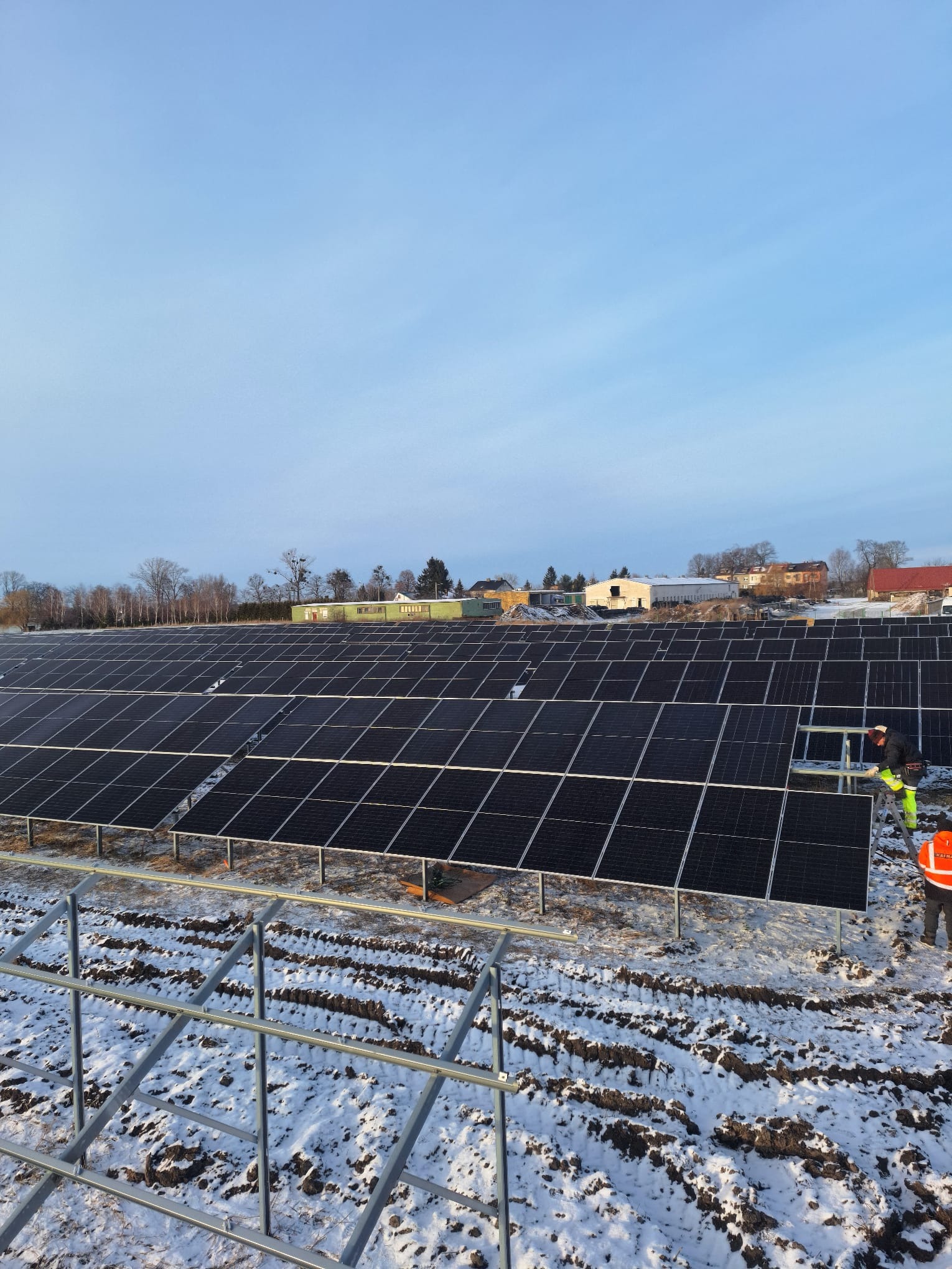 Rozległa farma paneli słonecznych w zimowym krajobrazie, z pracownikami w odblaskowych ubraniach podczas instalacji. Częściowo zasypana śniegiem ziemia.