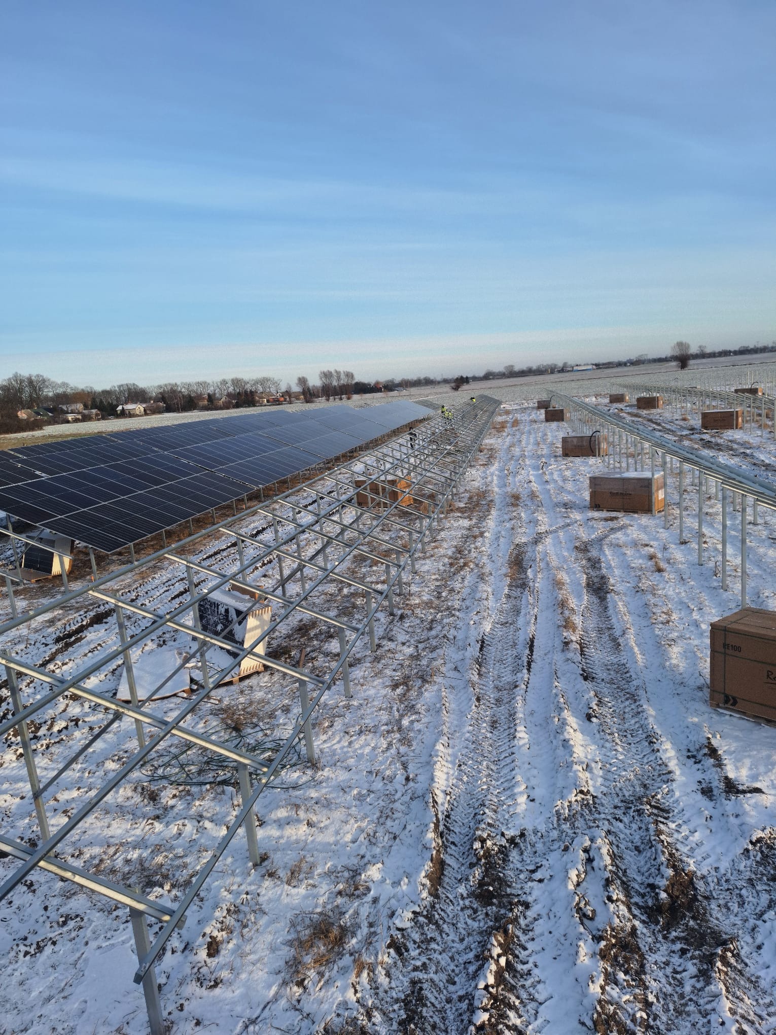 Montaż farmy fotowoltaicznej na zaśnieżonym terenie. Widoczne rzędy paneli i stalowa konstrukcja. Ślady opon na śniegu. Błękitne niebo w tle.