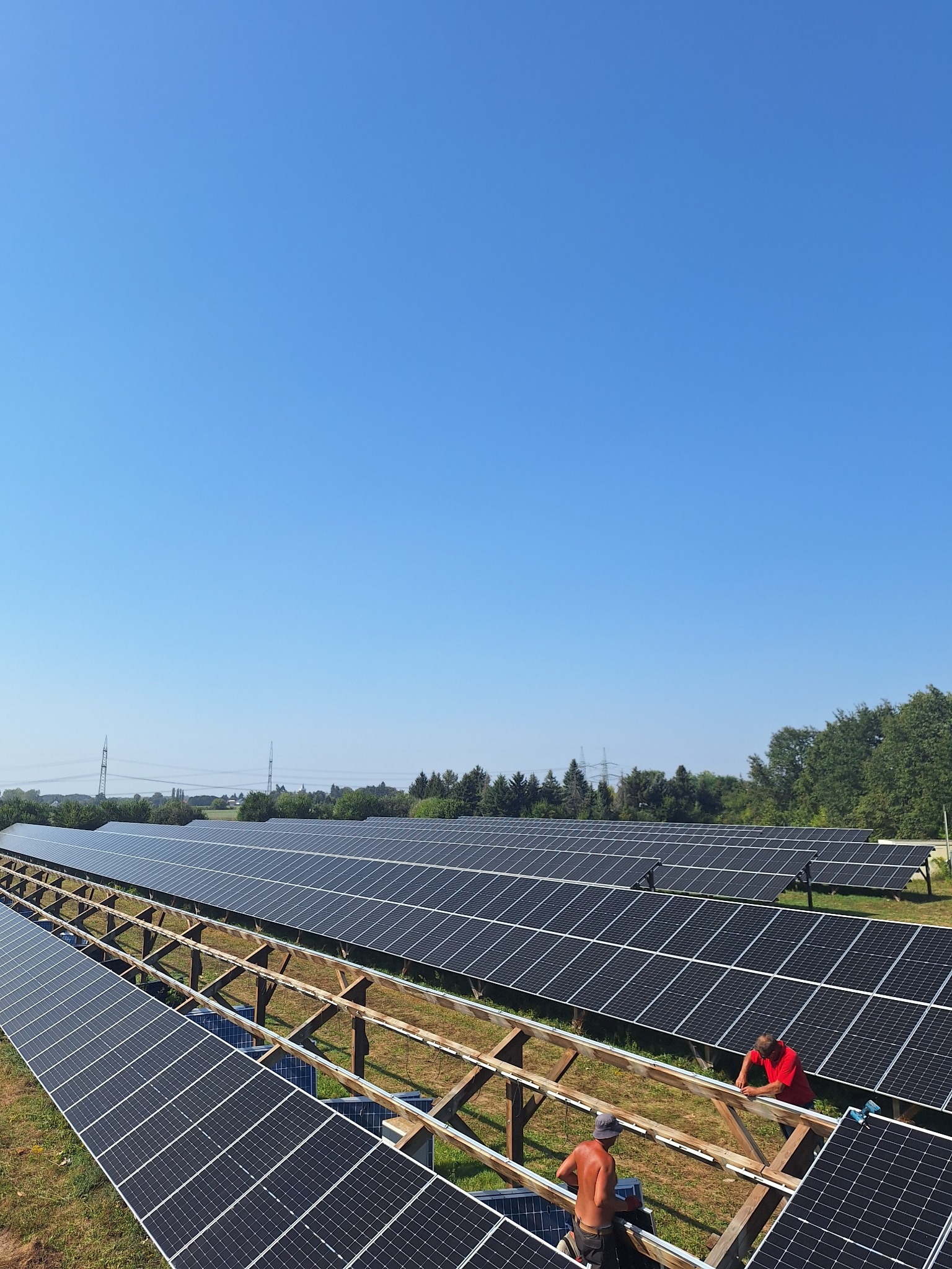 Instalacja paneli słonecznych na drewnianej konstrukcji przez dwóch monterów w słoneczny dzień. Widok na rozległą farmę fotowoltaiczną.
