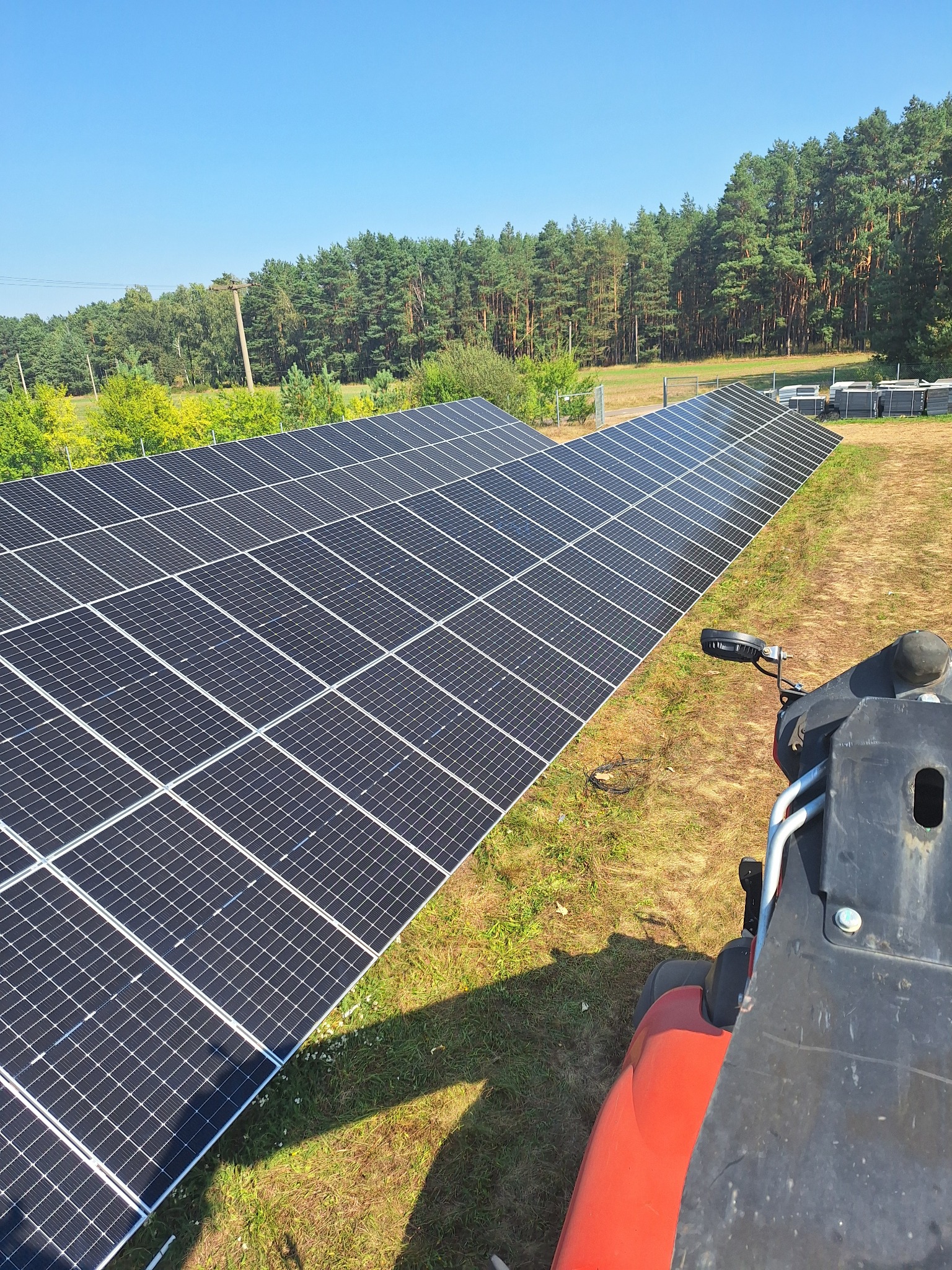 Rząd paneli słonecznych na trawiastym terenie z lasem w tle, widziany z perspektywy maszyny budowlanej. Błękitne niebo.