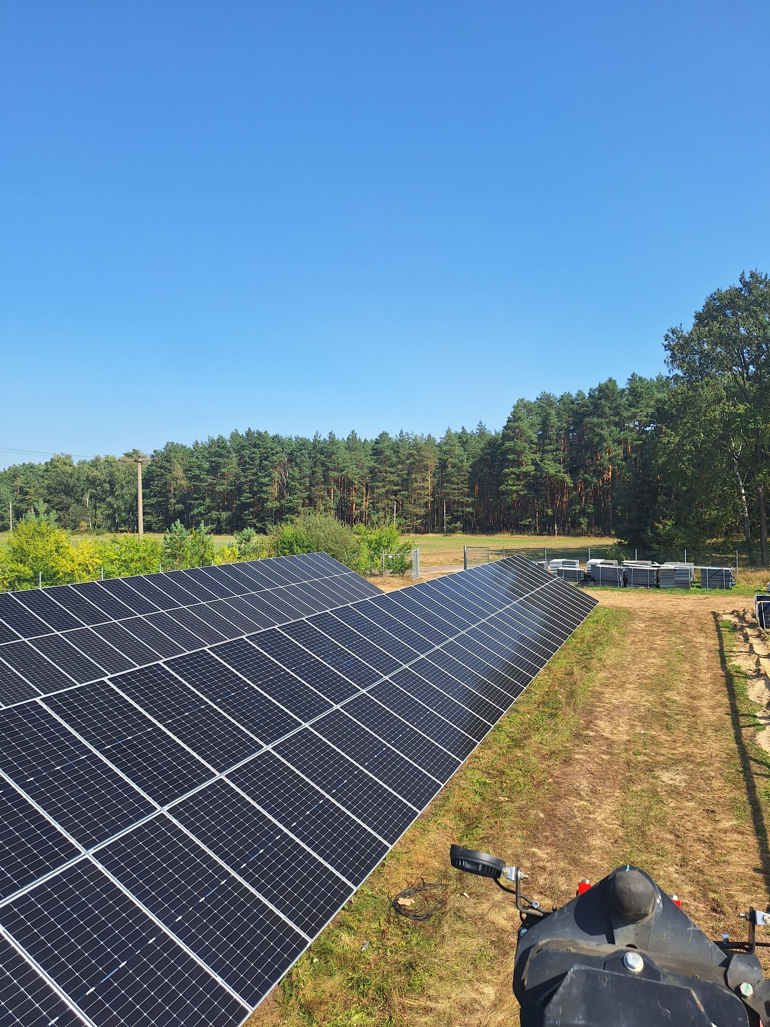 Rozległa instalacja paneli słonecznych na tle lasu i błękitnego nieba. Widok z góry, z elementami sprzętu montażowego w kadrze. Energia odnawialna, ekologiczne rozwiązanie.