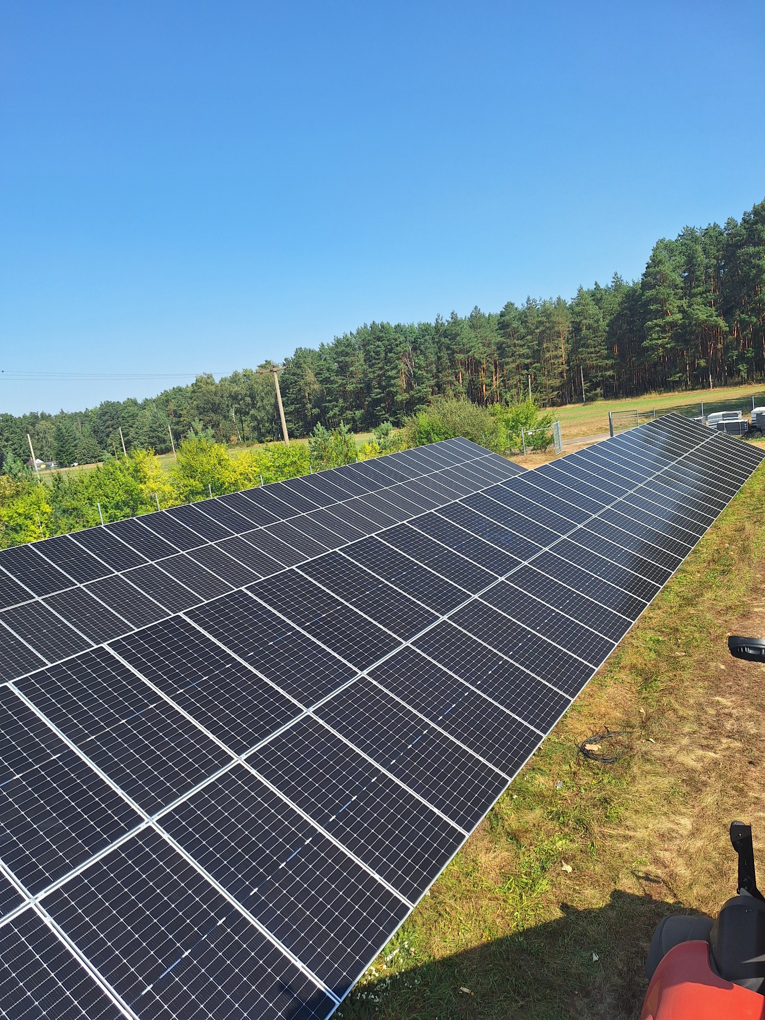 Rozległa instalacja fotowoltaiczna na zielonej trawie, z widokiem na las w tle, uchwycona w słoneczny dzień z perspektywy dachu, z fragmentem czerwonego traktora w kadrze.