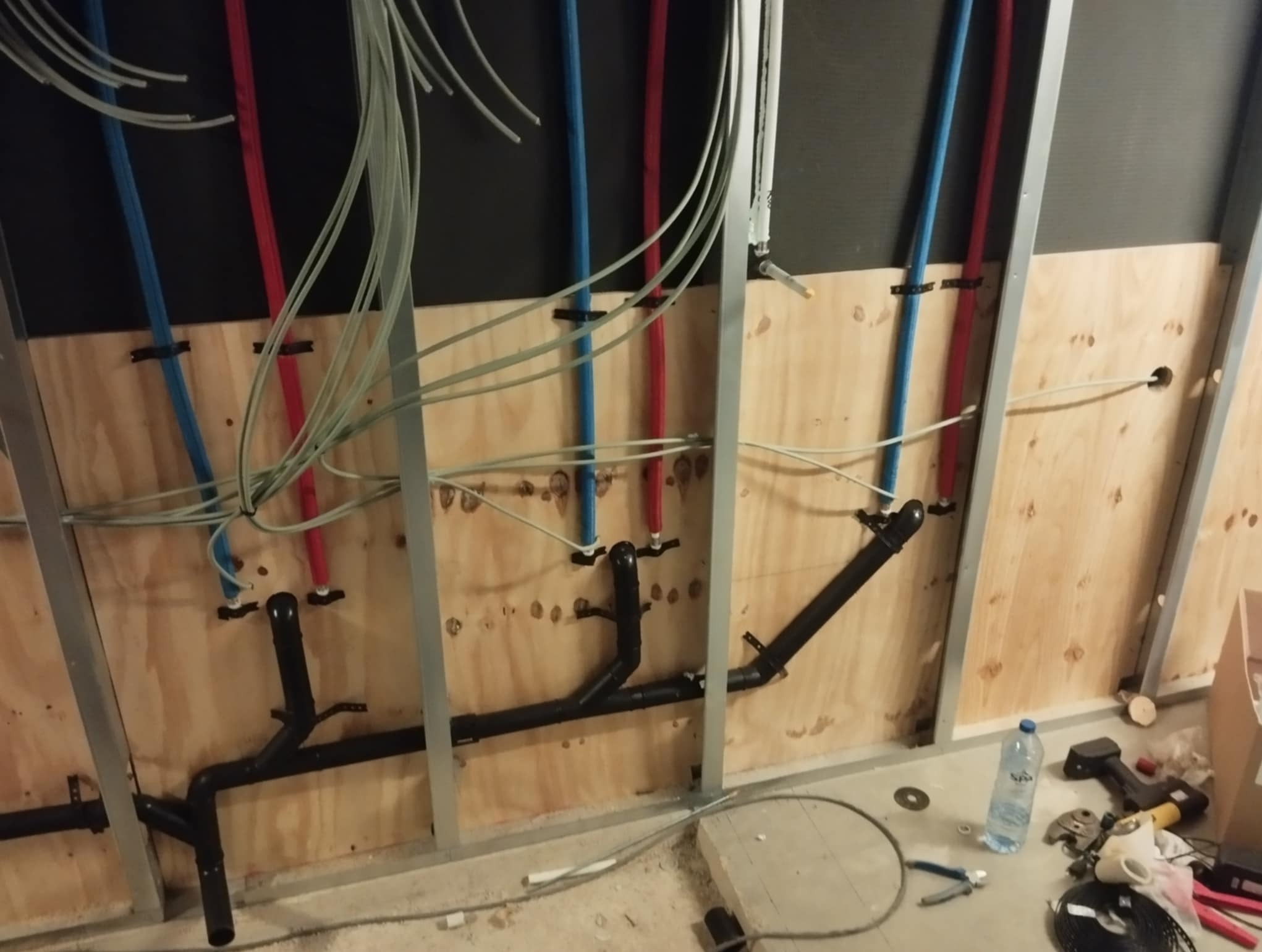 Instalacja wodno-kanalizacyjna wewnątrz suchej zabudowy, widoczne rury w kolorach niebieskim i czerwonym oraz czarne rury kanalizacyjne na drewnianej płycie.