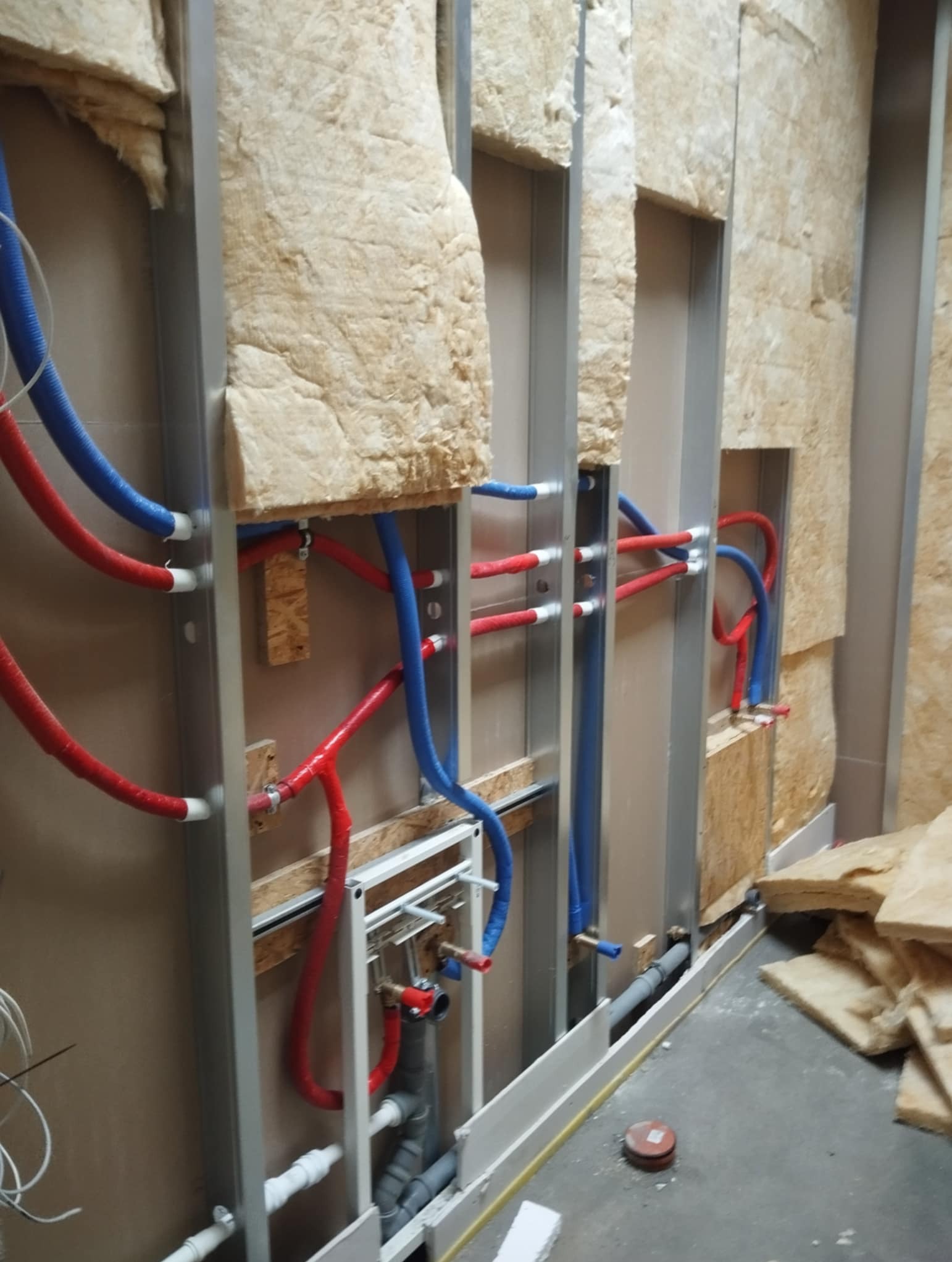 Wewnętrzna instalacja wodno-kanalizacyjna w zabudowie z izolacją, rury czerwone i niebieskie, metalowy stelaż. Widoczne elementy montażowe i izolacja.