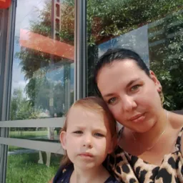 Kobieta w bluzce w panterkę i mała dziewczynka siedzą na przystanku autobusowym z widokiem na zieleń. Przezroczyste szyby odbijają niebo i drzewa.