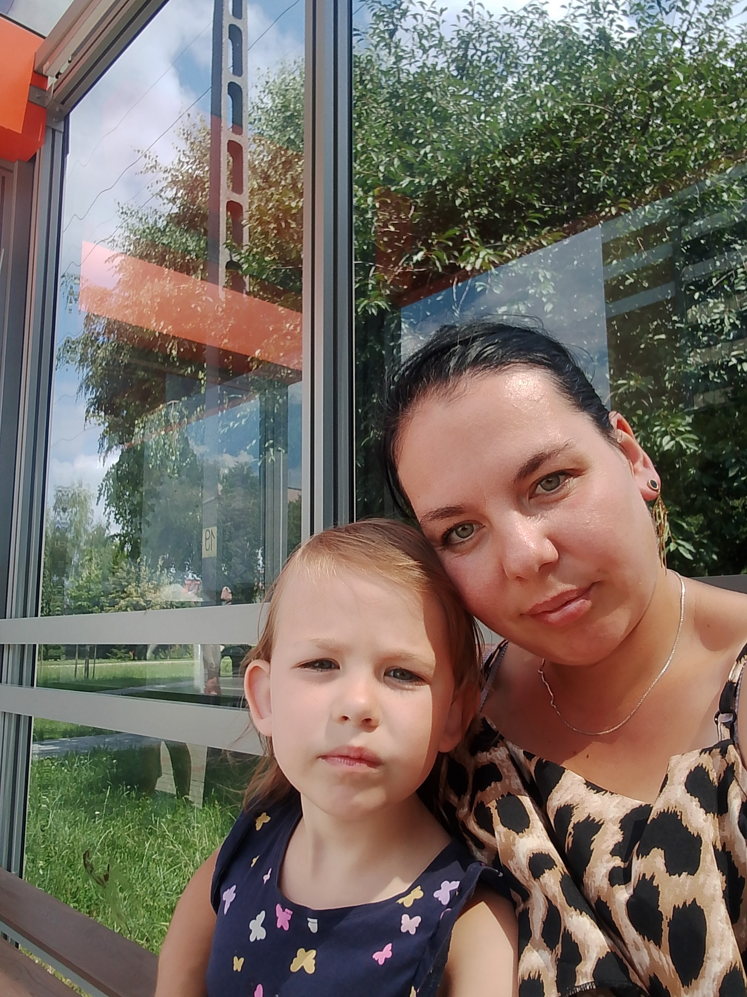 Kobieta w bluzce w panterkę i mała dziewczynka siedzą na przystanku autobusowym z widokiem na zieleń. Przezroczyste szyby odbijają niebo i drzewa.